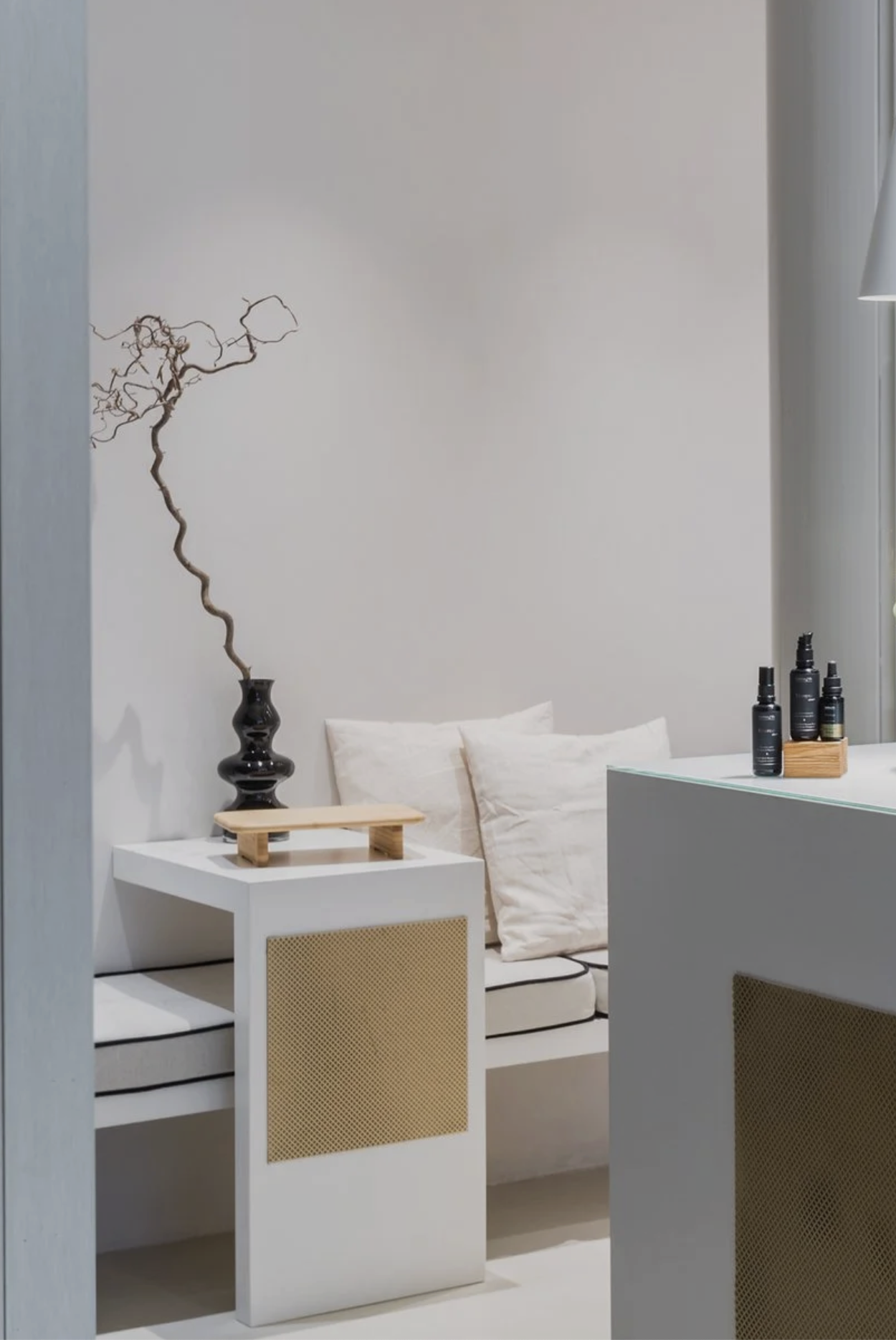Minimalist interior with a white bench with black piping, beige pillows, a white side table with a black vase and dried twigs, and bottles of skincare on a counter.
