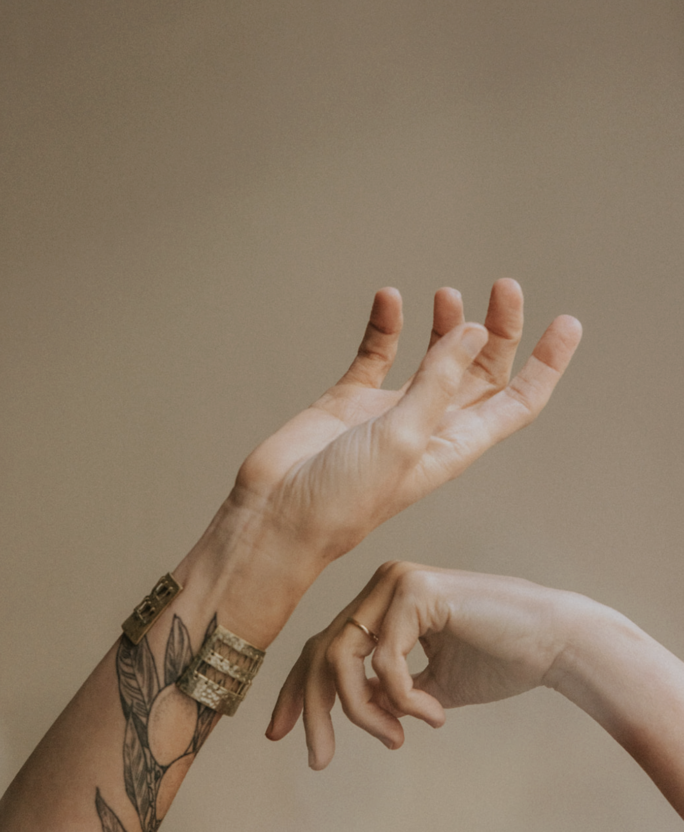 Two hands, one with a tattoo and bracelets reaching upward, the other with a ring on the finger and making a gesture, against a plain background.