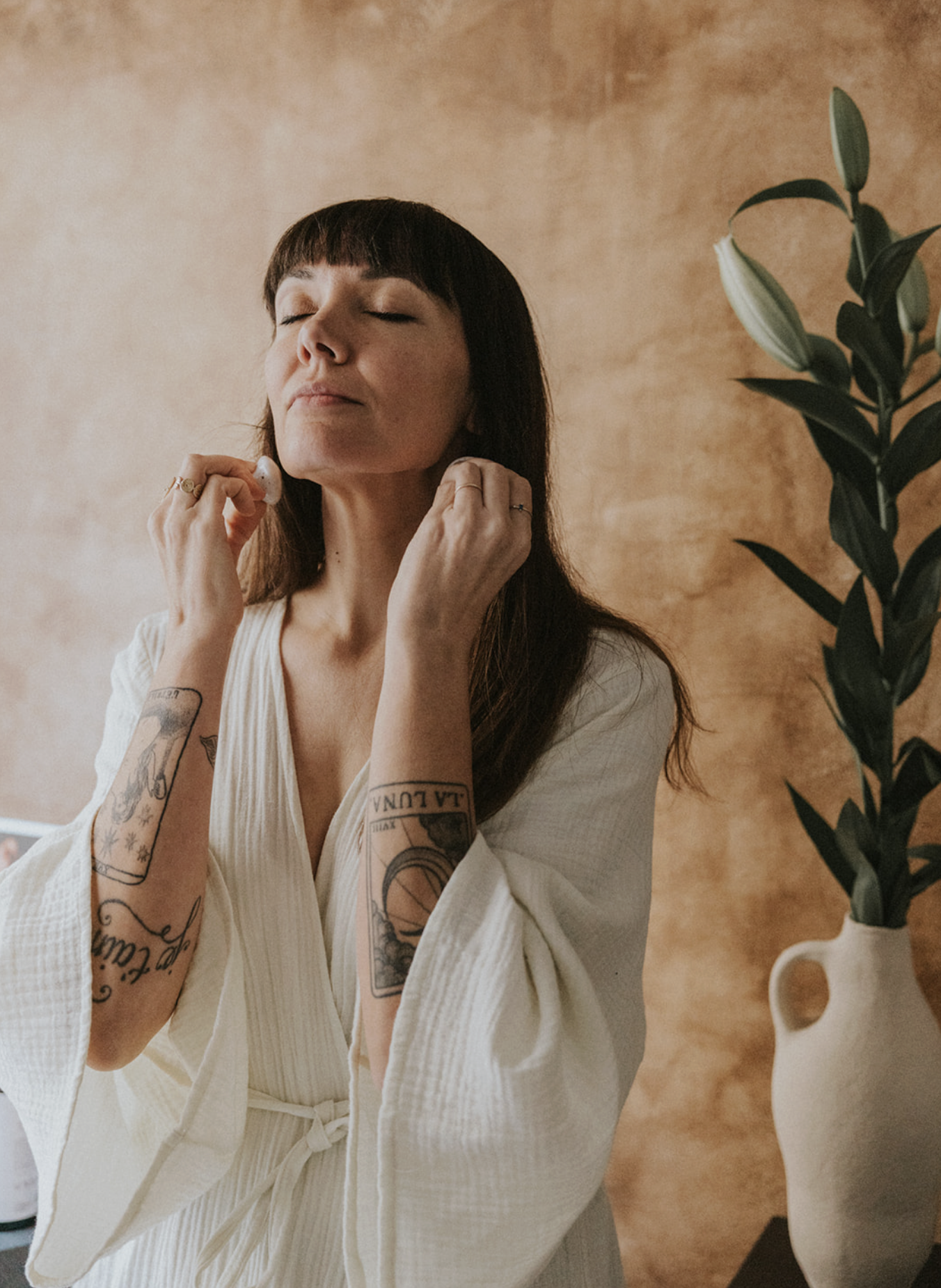 A woman with dark brown hair and tattoos on her arms, wearing a white robe, is relaxing with her eyes closed and hands gently touching her neck in a serene indoor setting with a potted plant and a warm-toned background.