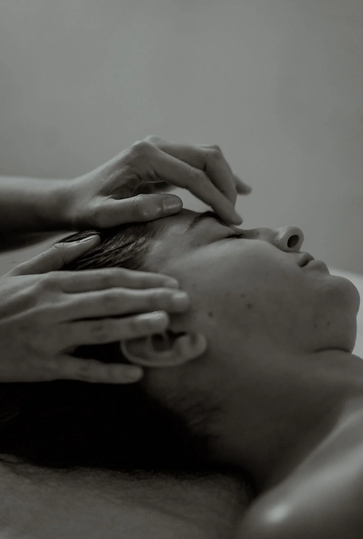 Close-up black and white photo of a woman lying down with eyes closed, while two hands gently touch her forehead and temple.