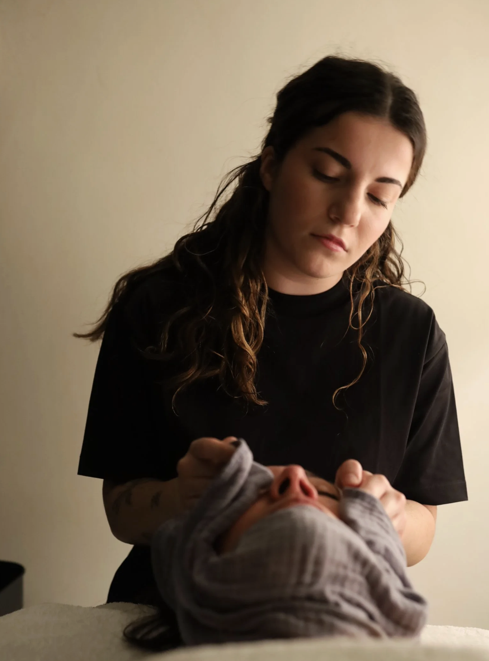 A person receiving a facial massage from a woman in a black shirt, with a neutral-colored wall in the background.