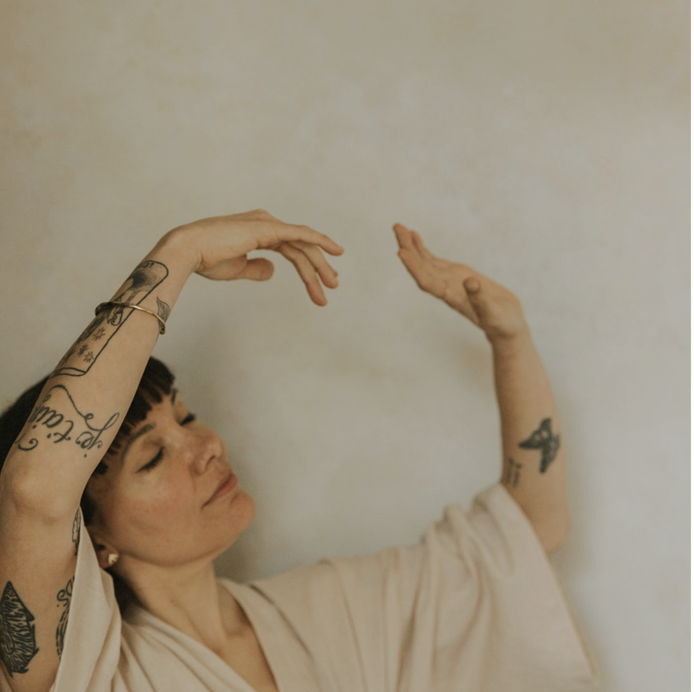 A woman with tattoos on her arms and short dark hair, closing her eyes with a peaceful expression, raising her hands gracefully near her head against a plain beige wall.