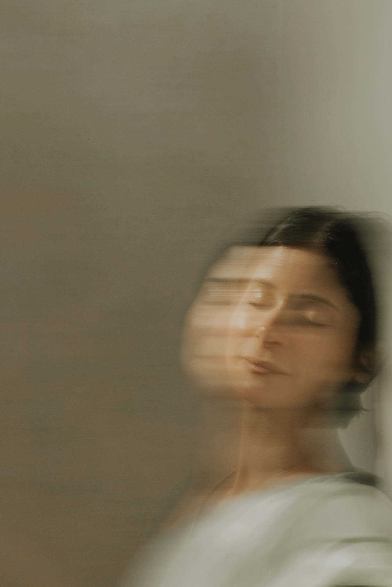 A woman with short dark hair and light skin, with a blurred, motion effect on her face, standing against a plain wall.