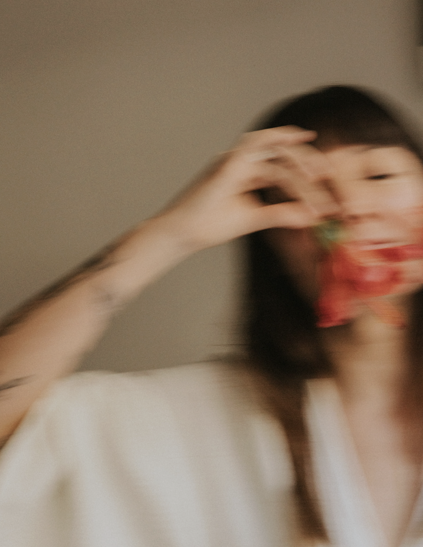 Blurry photo of a woman with dark hair wearing a white top, making a hand gesture near her eye and smiling.
