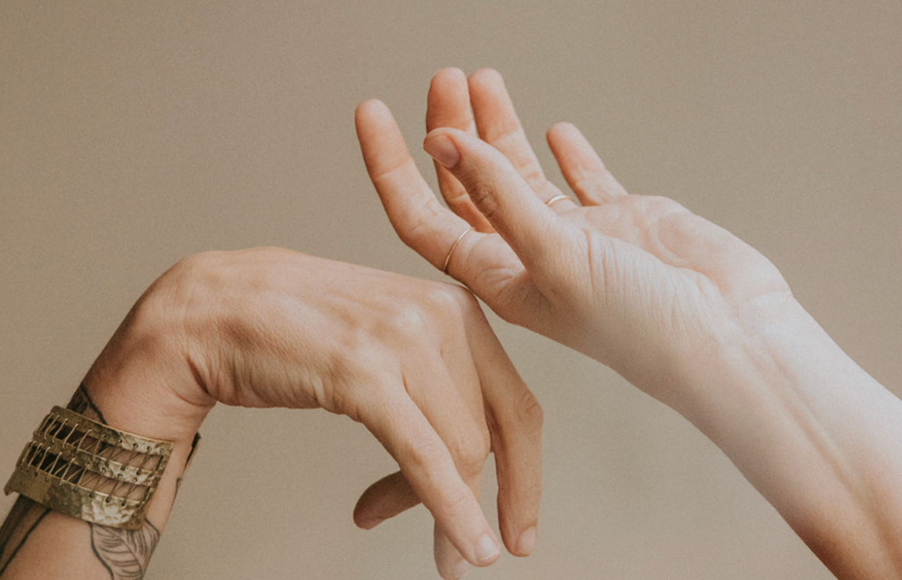 Two hands, one with a tattoo and a gold watch, and the other with thin rings, touching fingers against a neutral background.