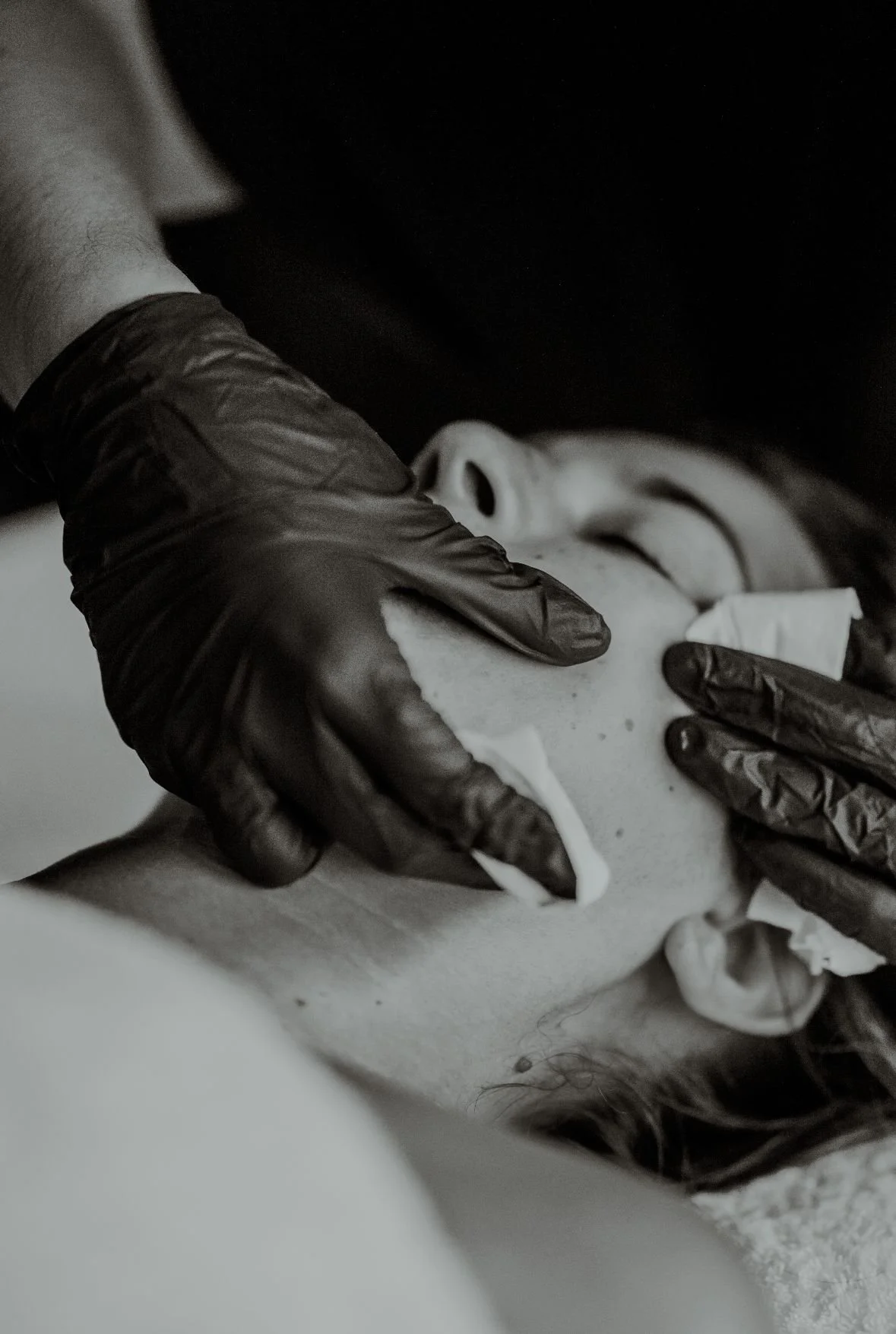 A person wearing black gloves is applying a facial mask to another person's face, who is lying down with eyes closed.