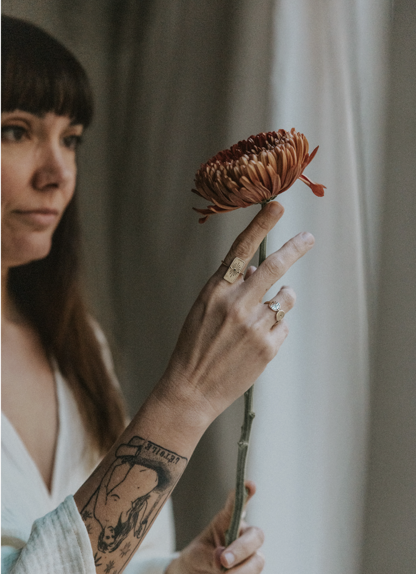 A woman with tattoos on her arm holding a flower in front of a window.