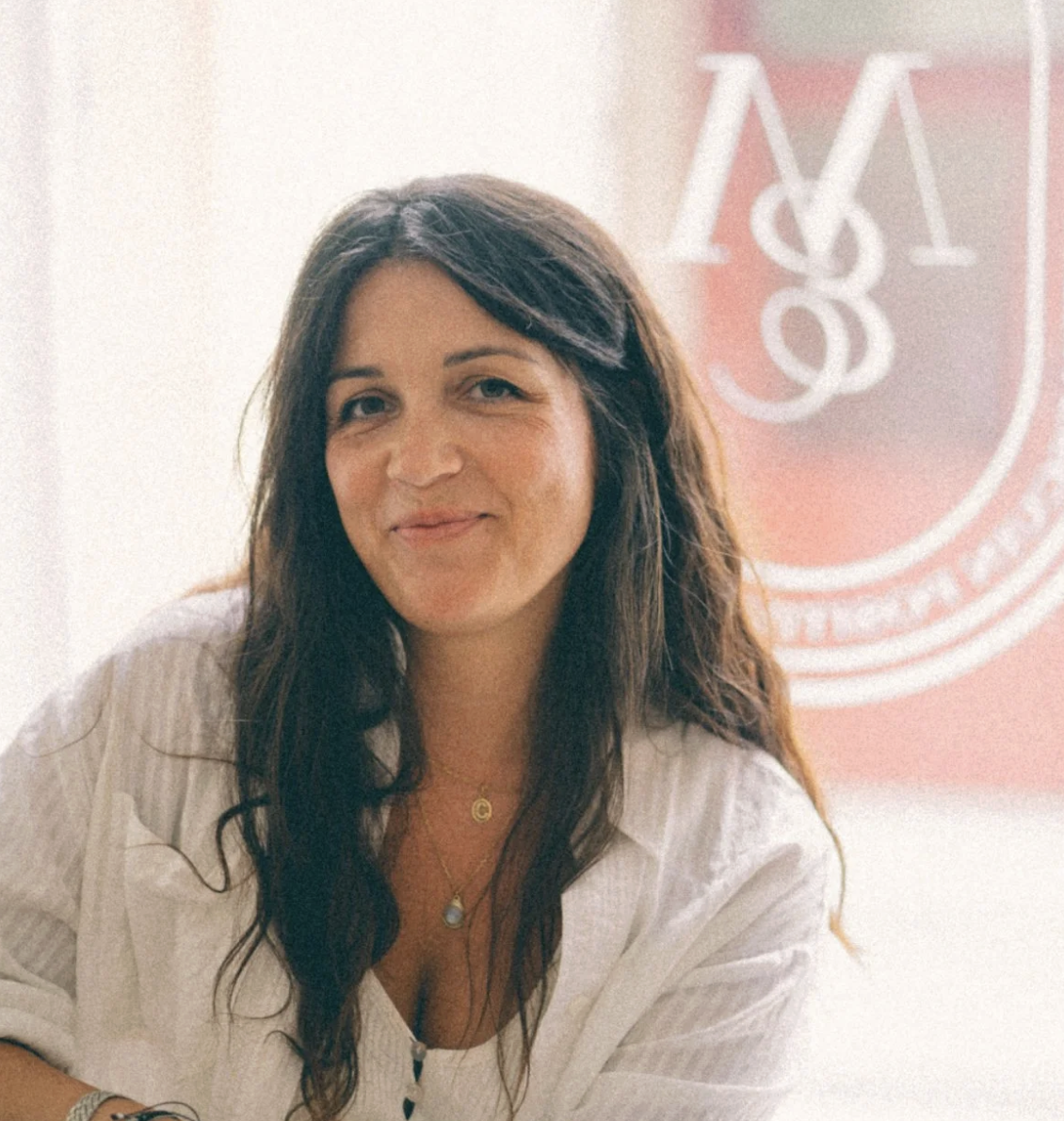 A woman with long brown hair, wearing a white shirt and layered necklaces, smiling in front of a blurred background with a partial logo or emblem.