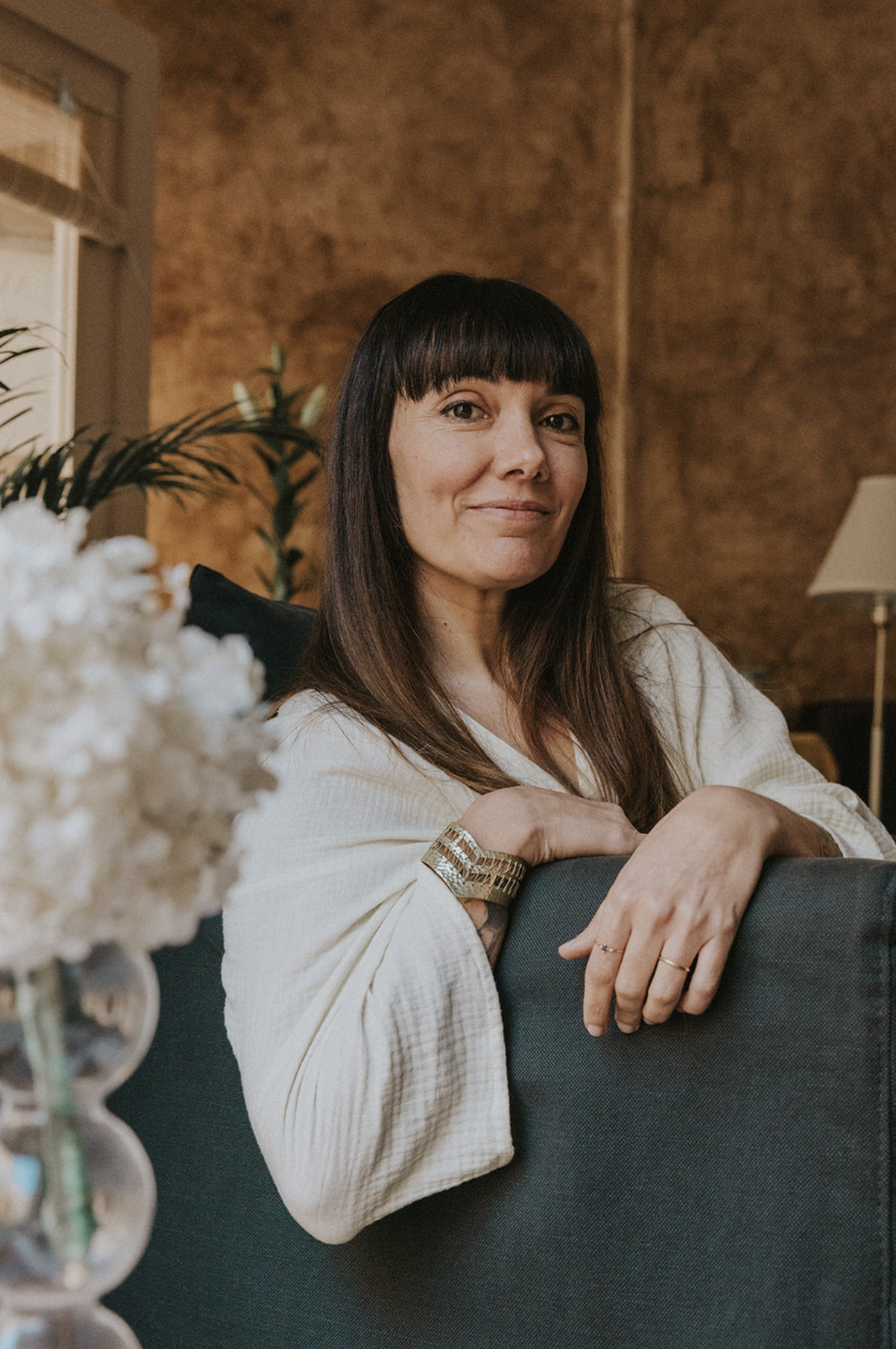 Woman with dark hair and bangs sitting on a sofa, smiling, in a cozy room with brown walls, a lamp, and houseplants, with a blurred white flower arrangement in the foreground.