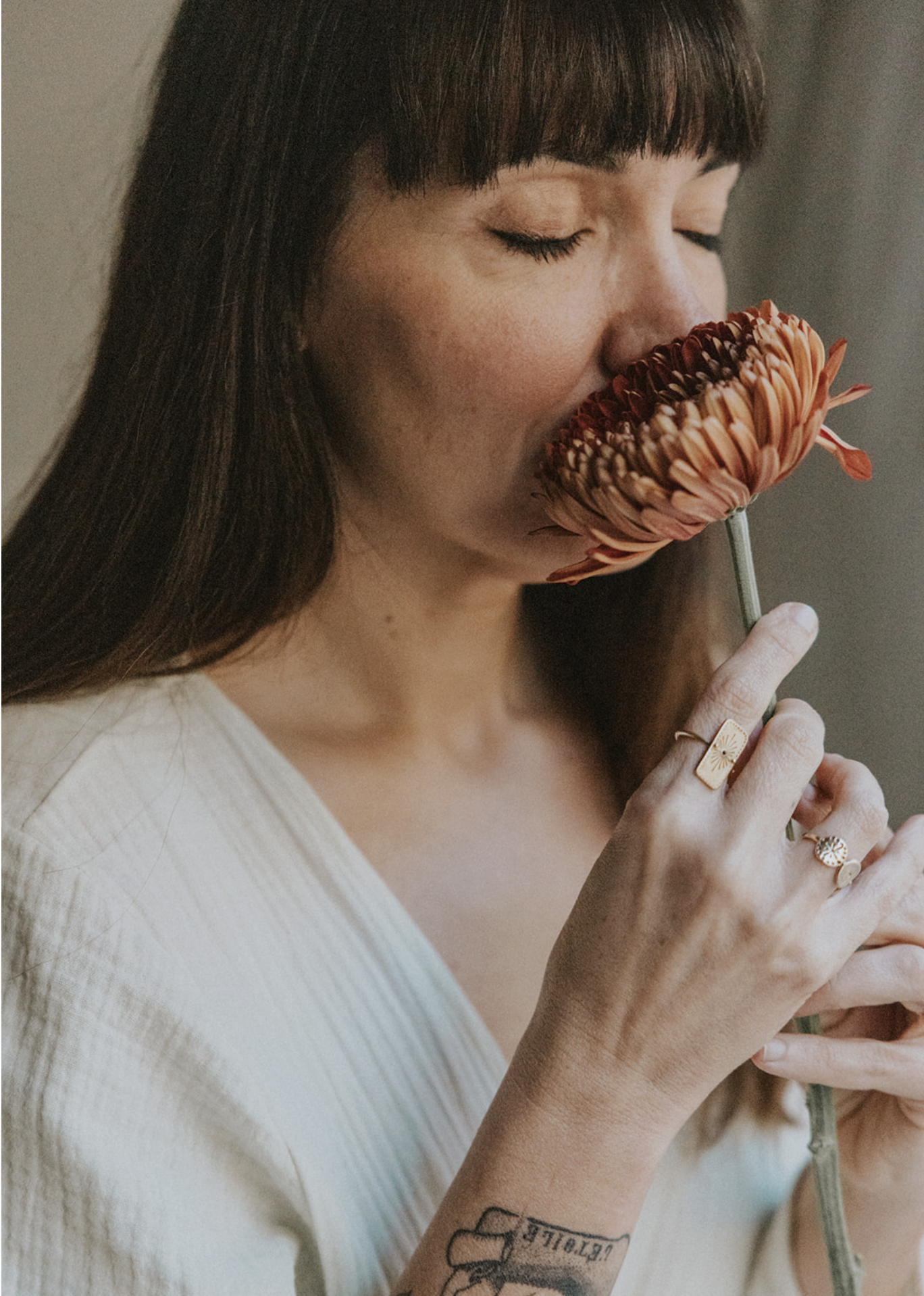 A woman with long brown hair and bangs holds a large flower close to her face, with her eyes closed.