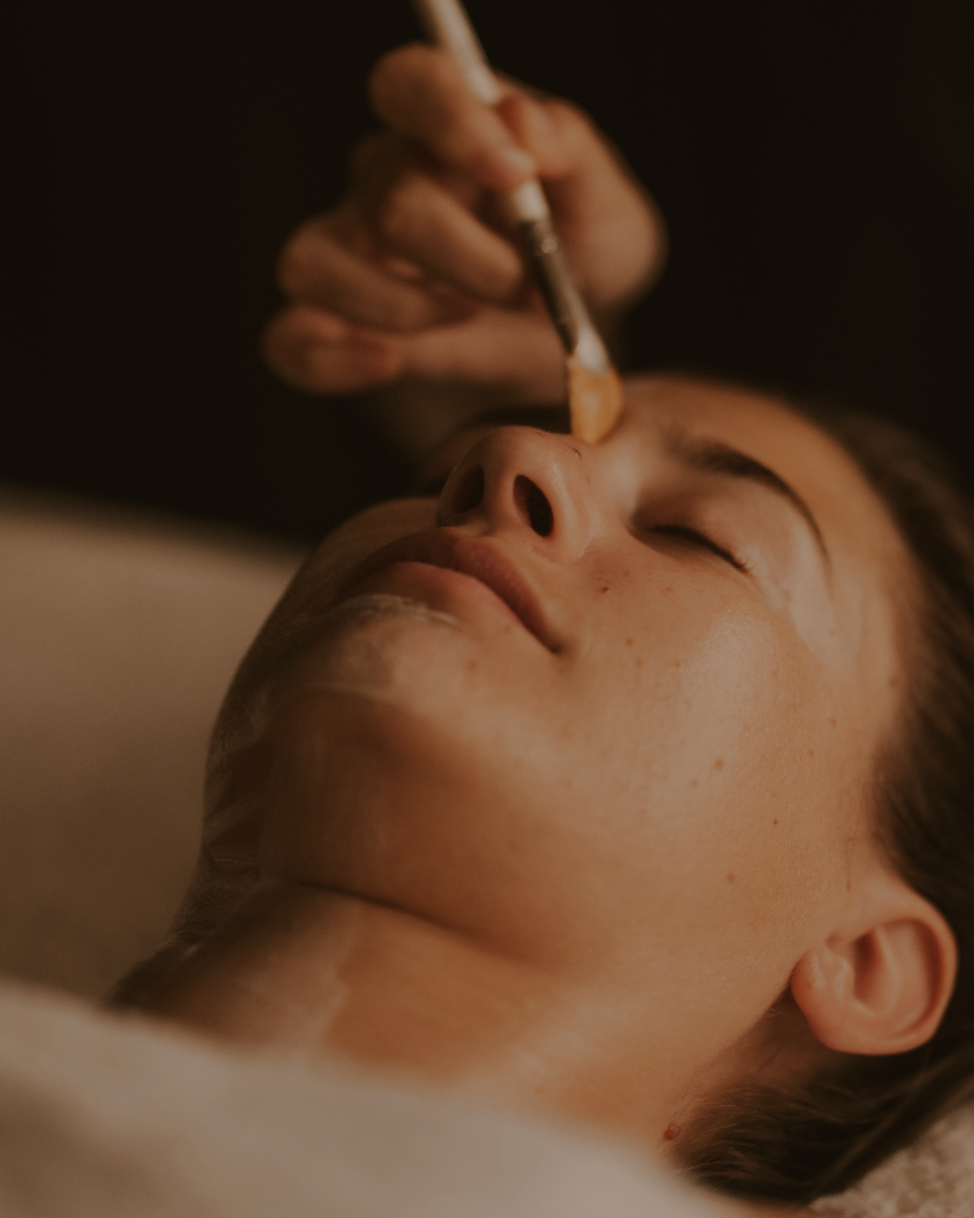 A woman lying down with her eyes closed, receiving a facial treatment as a professional applies product with a brush to her face.