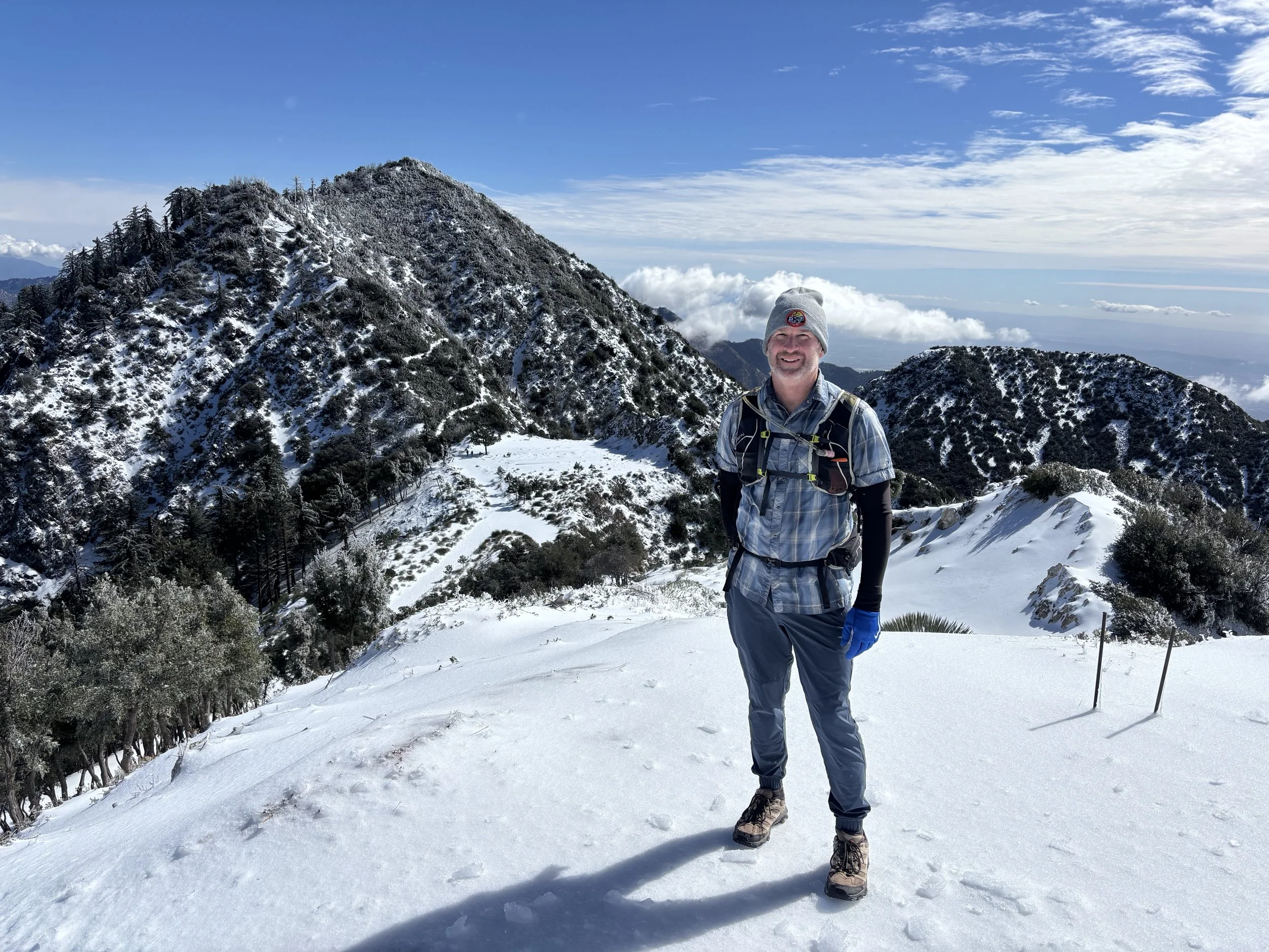 A man hiking on snowy mountain terrain during daytime, wearing a gray beanie, plaid shirt, gray pants, hiking boots, and gloves, with a backpack and trekking poles, surrounded by snow-covered trees and mountain peaks under a partly cloudy sky.