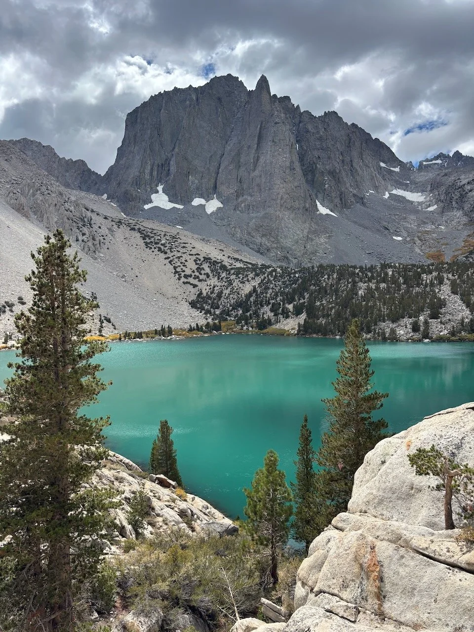 Turquoise alpine lake at Big Pine Lakes in the Eastern Sierras, surrounded by pine trees and rocky mountain terrain under a cloudy sky.