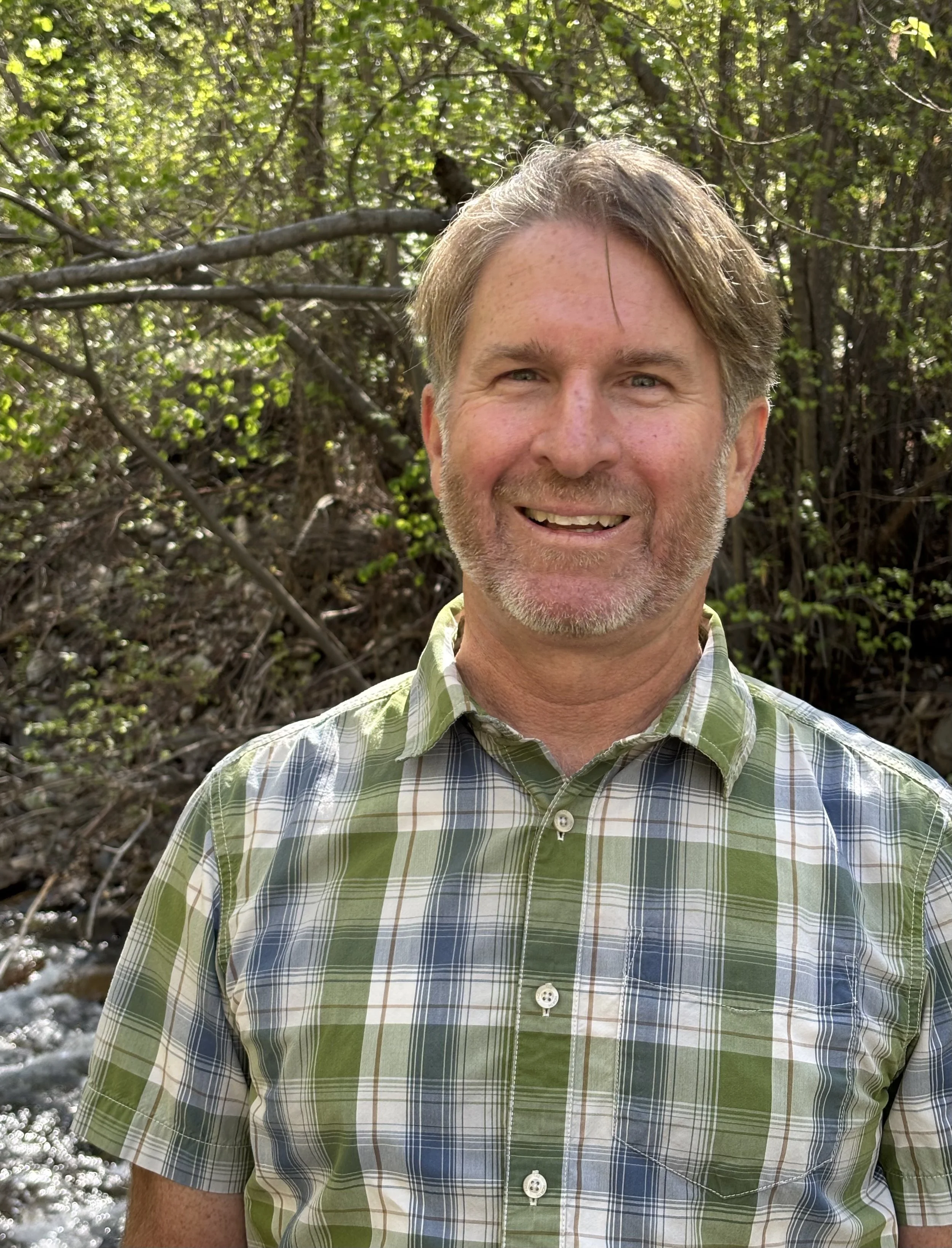 A close-up of a smiling man with gray hair and a beard, wearing a green, gray, and white plaid shirt, standing outdoors in front of a dense, green forest with sunlight filtering through the leaves.