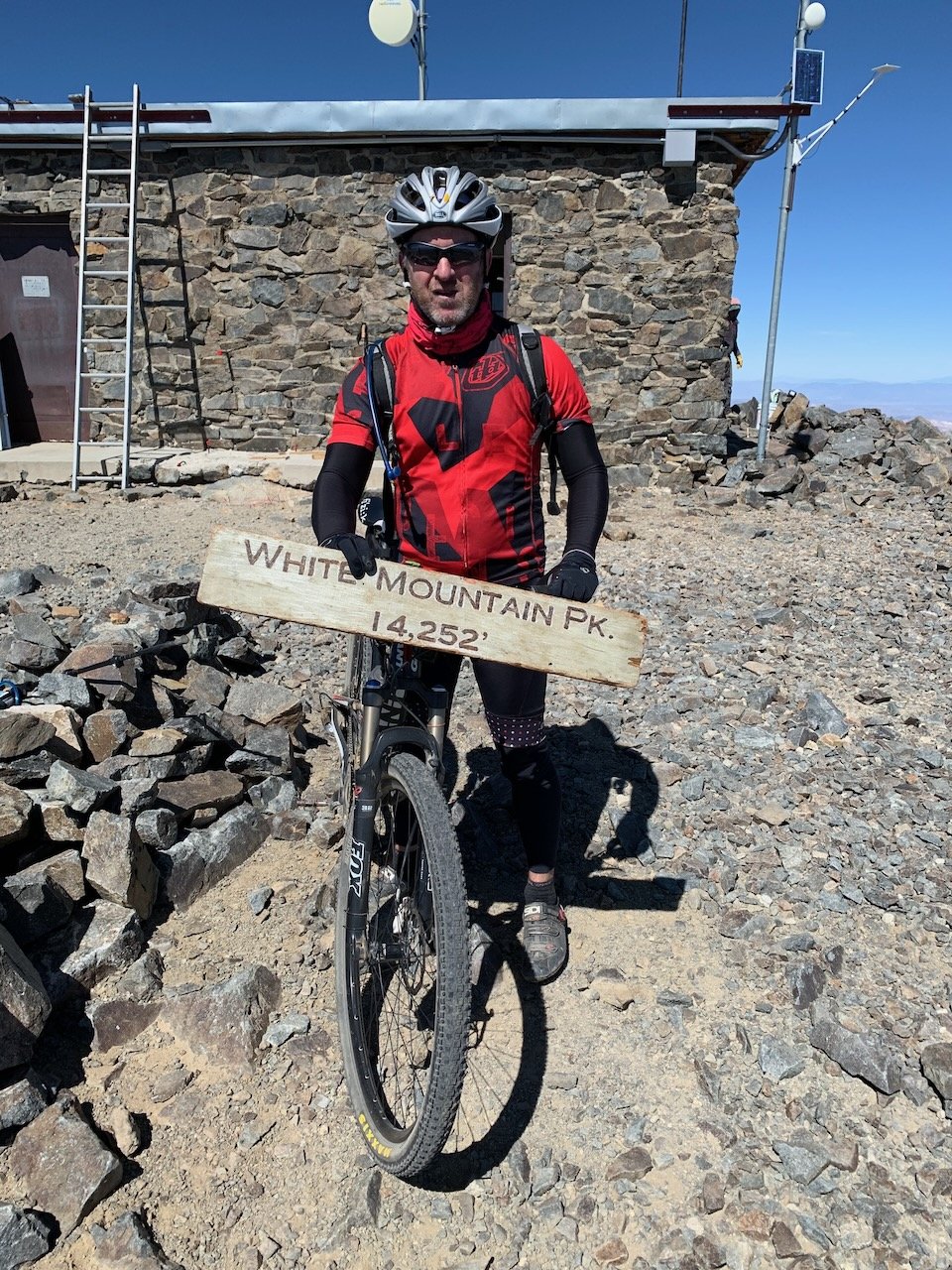 Ian Tervet mountain biking to the summit of White Mountain Peak (14,252') above Bishop, California, standing beside the summit sign in cycling gear near the historic research station and antennas.