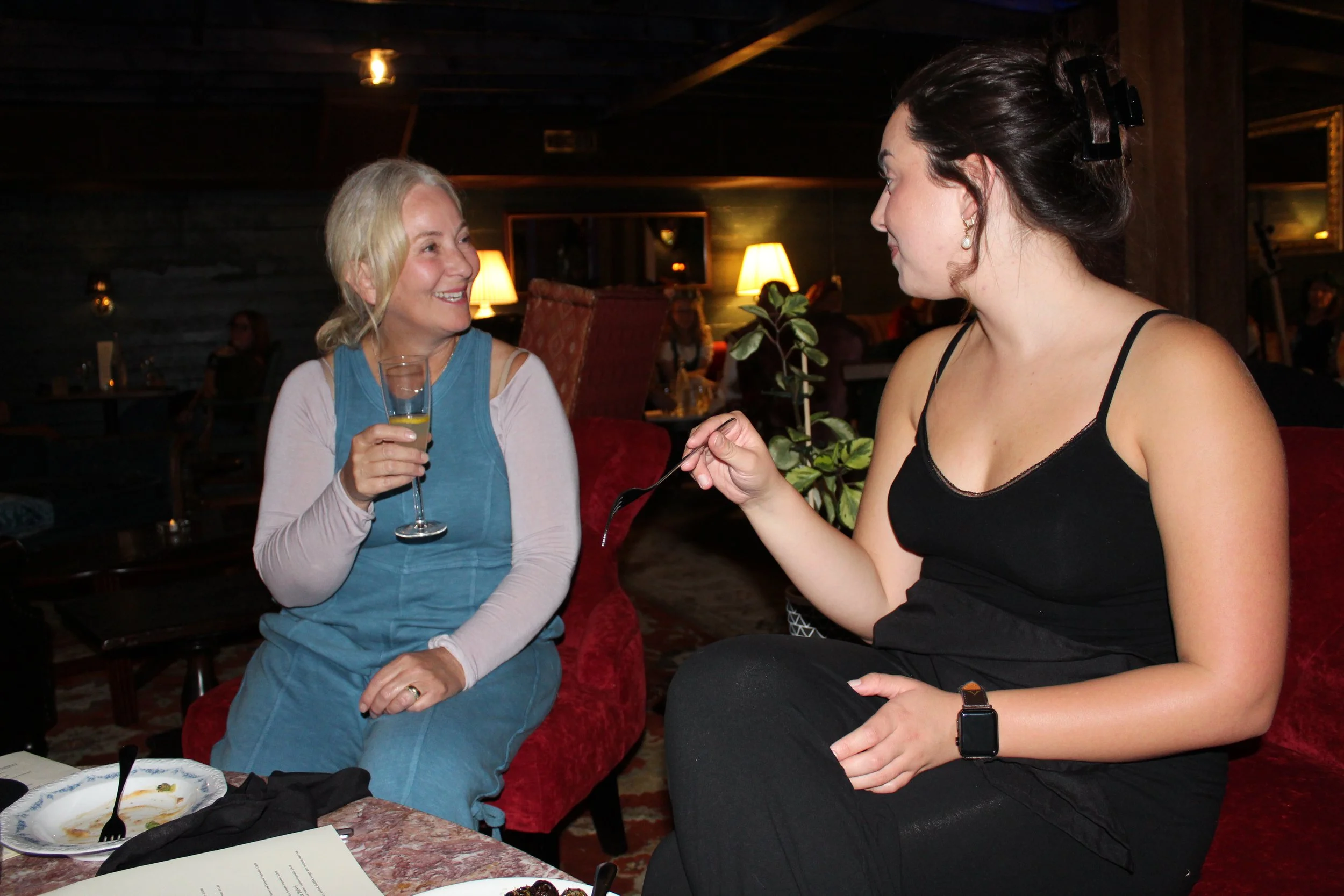 Two women sitting at a restaurant table. The woman on the left has blonde hair and is holding a glass of champagne, smiling at the woman on the right. The woman on the right has dark hair tied back, is wearing a black top, and is holding a fork. Ther