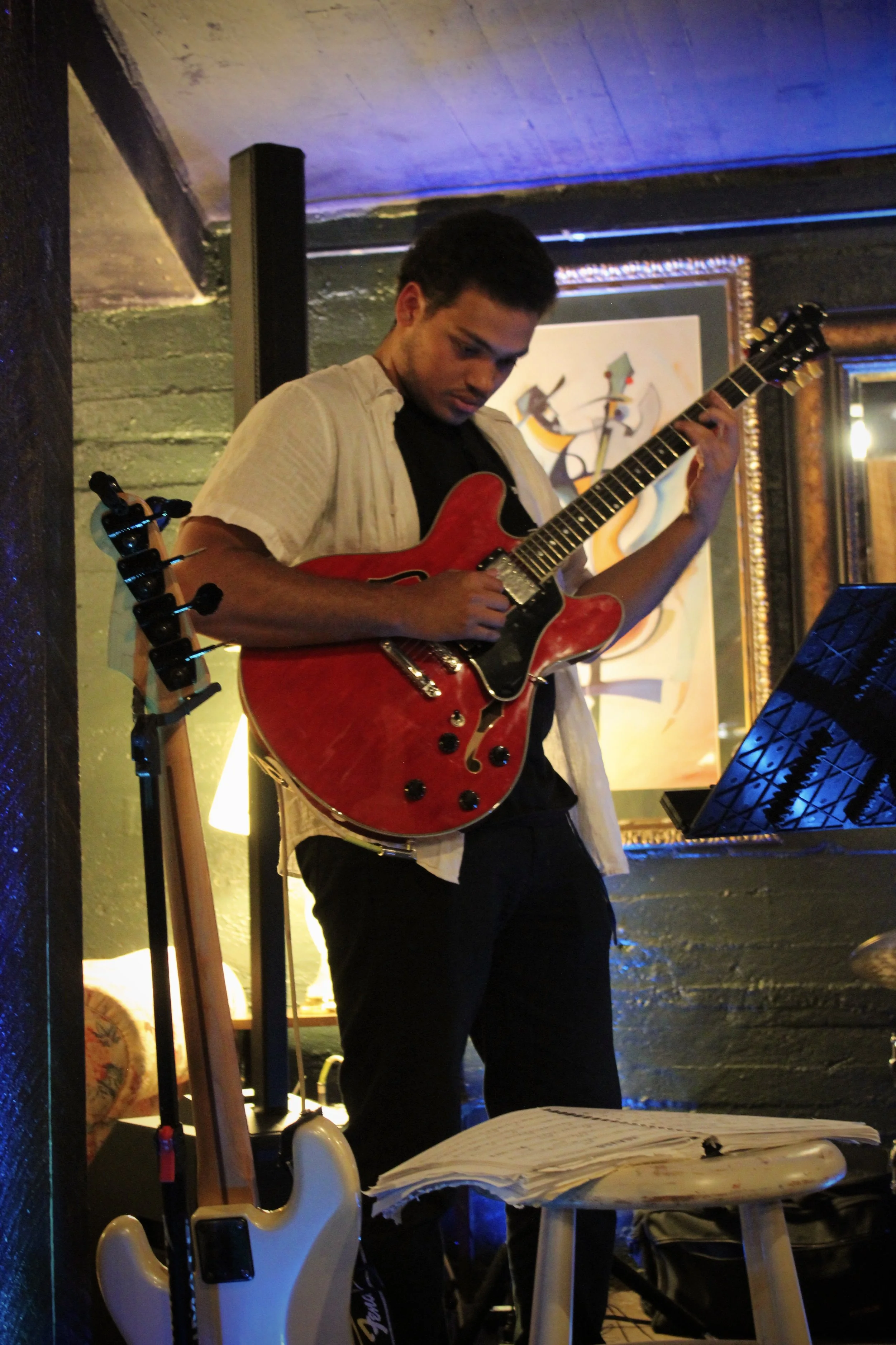 A young man playing an electric guitar on stage in a dimly lit room, with piano music sheets on a small table in the foreground and artwork hanging on the wall in the background.