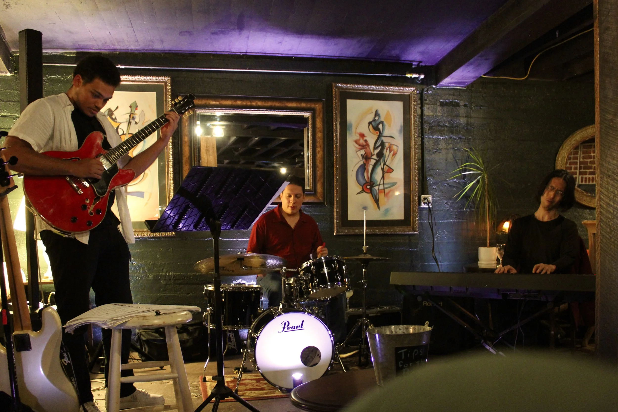 A jazz band performing in a cozy, intimate venue. The band includes a guitarist, a drummer, and a pianist. The venue features dark walls, framed abstract art, and subdued lighting with purple accents.