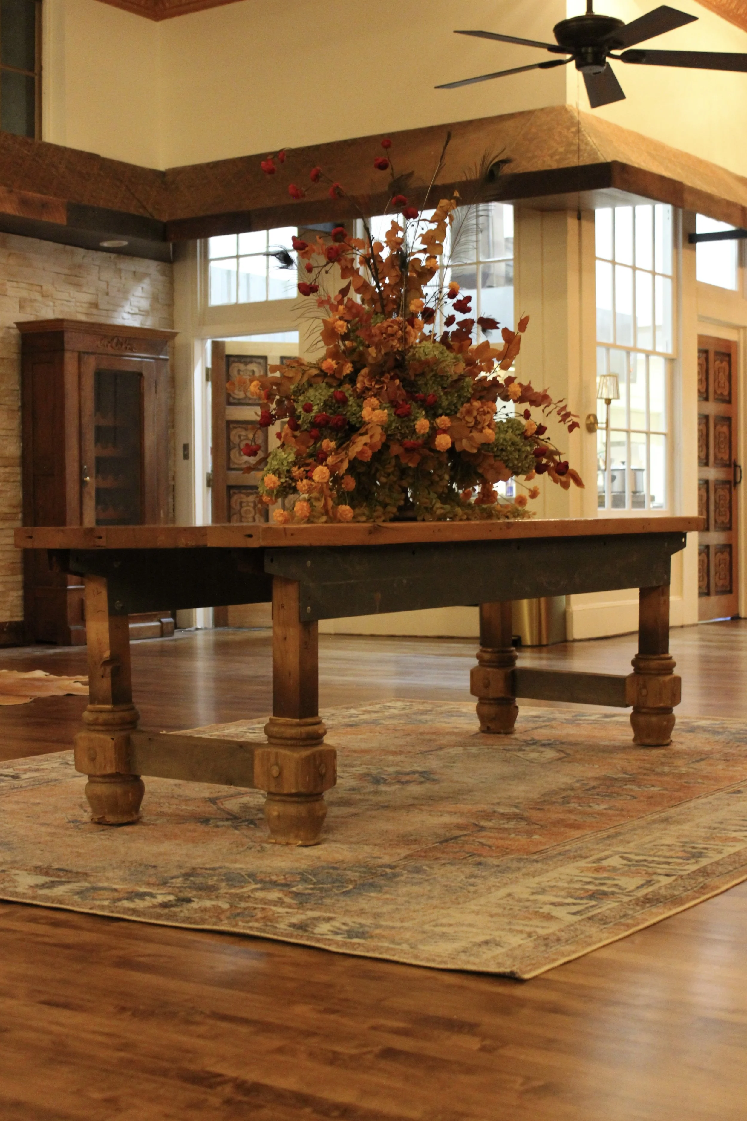 A wooden table with a large autumn flower arrangement in a vase, placed on a patterned rug in a cozy living room with large windows and a ceiling fan.