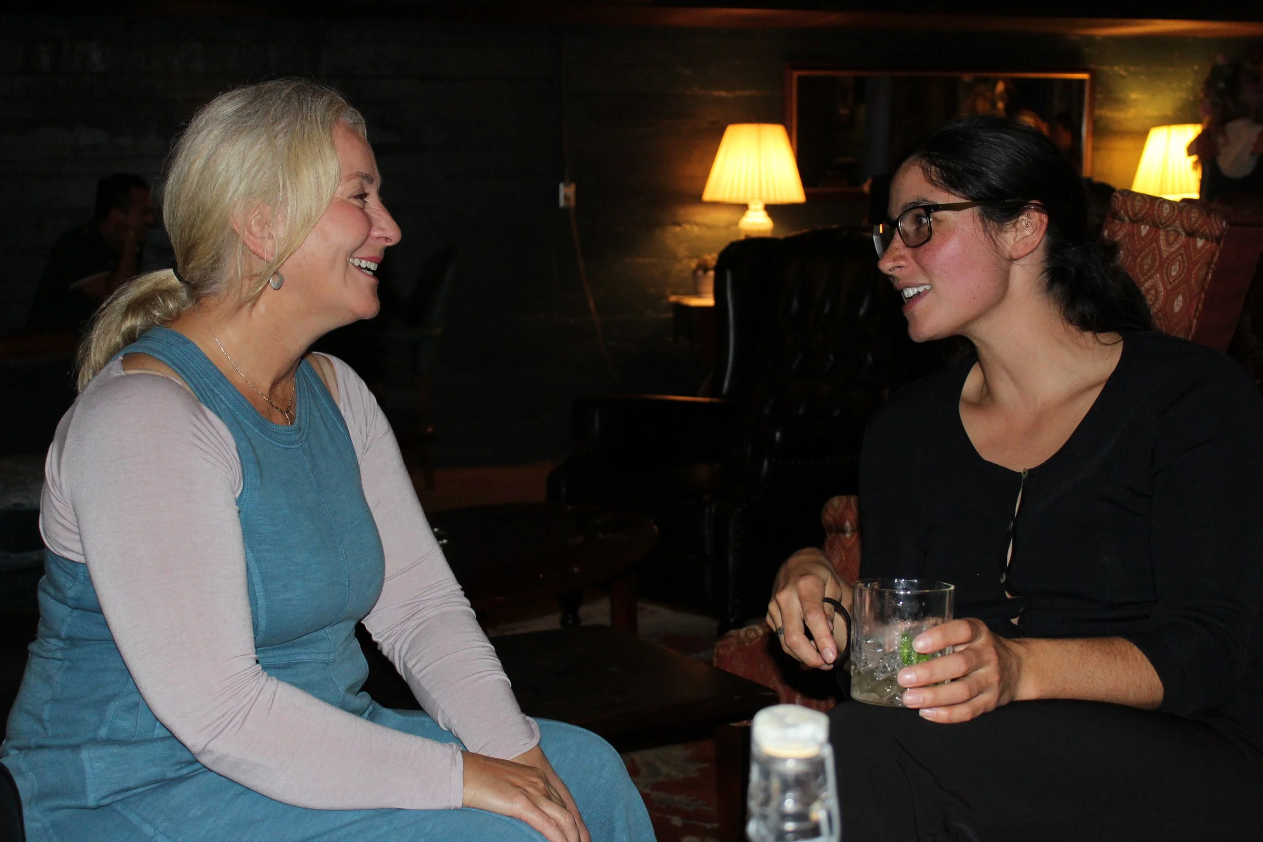 Two women smiling and talking in a dimly lit room with warm lamp lighting, one of them holding a drink.