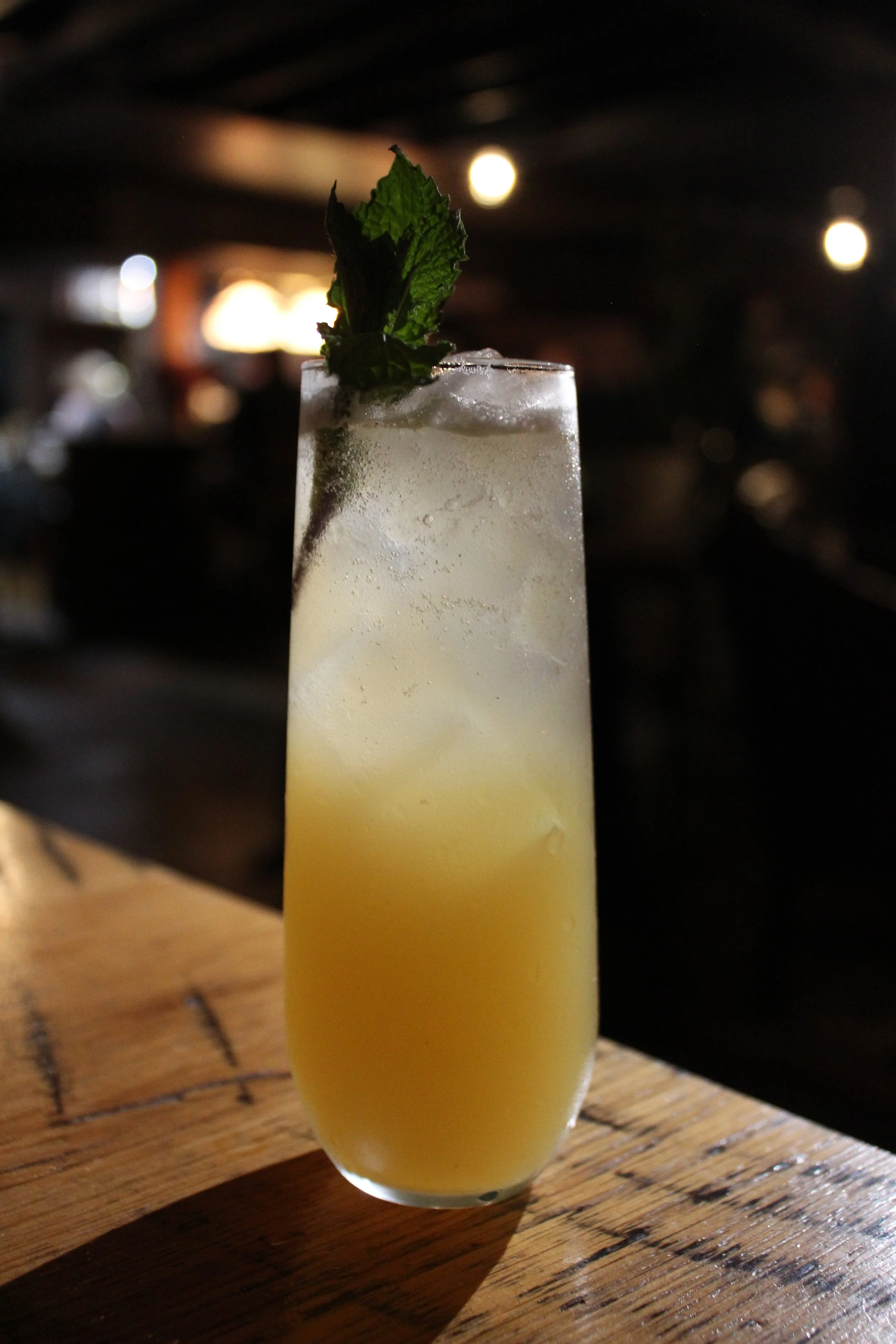 A tall glass filled with a yellowish beverage, ice cubes, and garnished with fresh mint leaves, set on a wooden surface with a dark, blurred background.