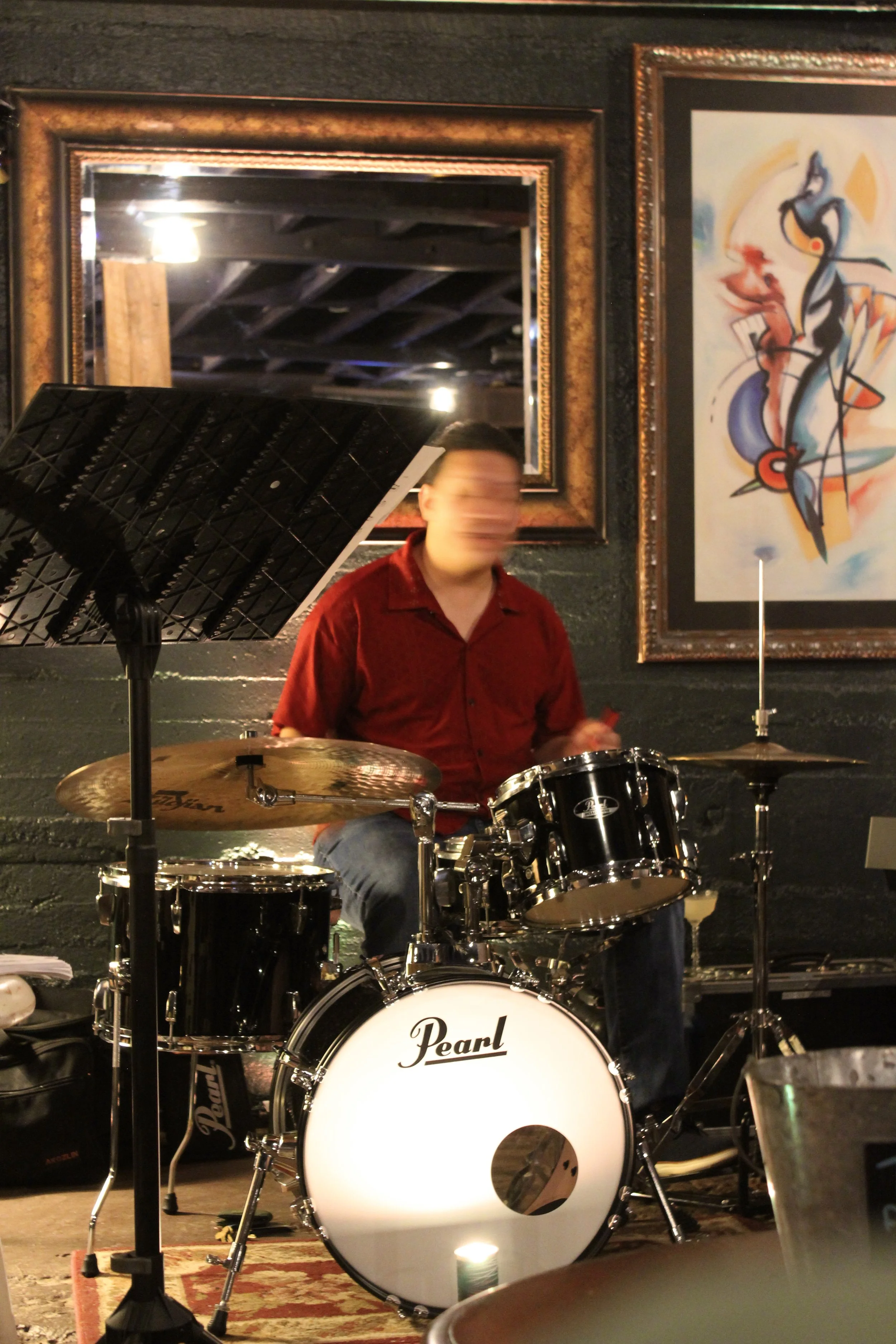 A person playing a drum set with a Pearl bass drum, in a room with dark walls and framed artwork, possibly during a musical performance or practice.