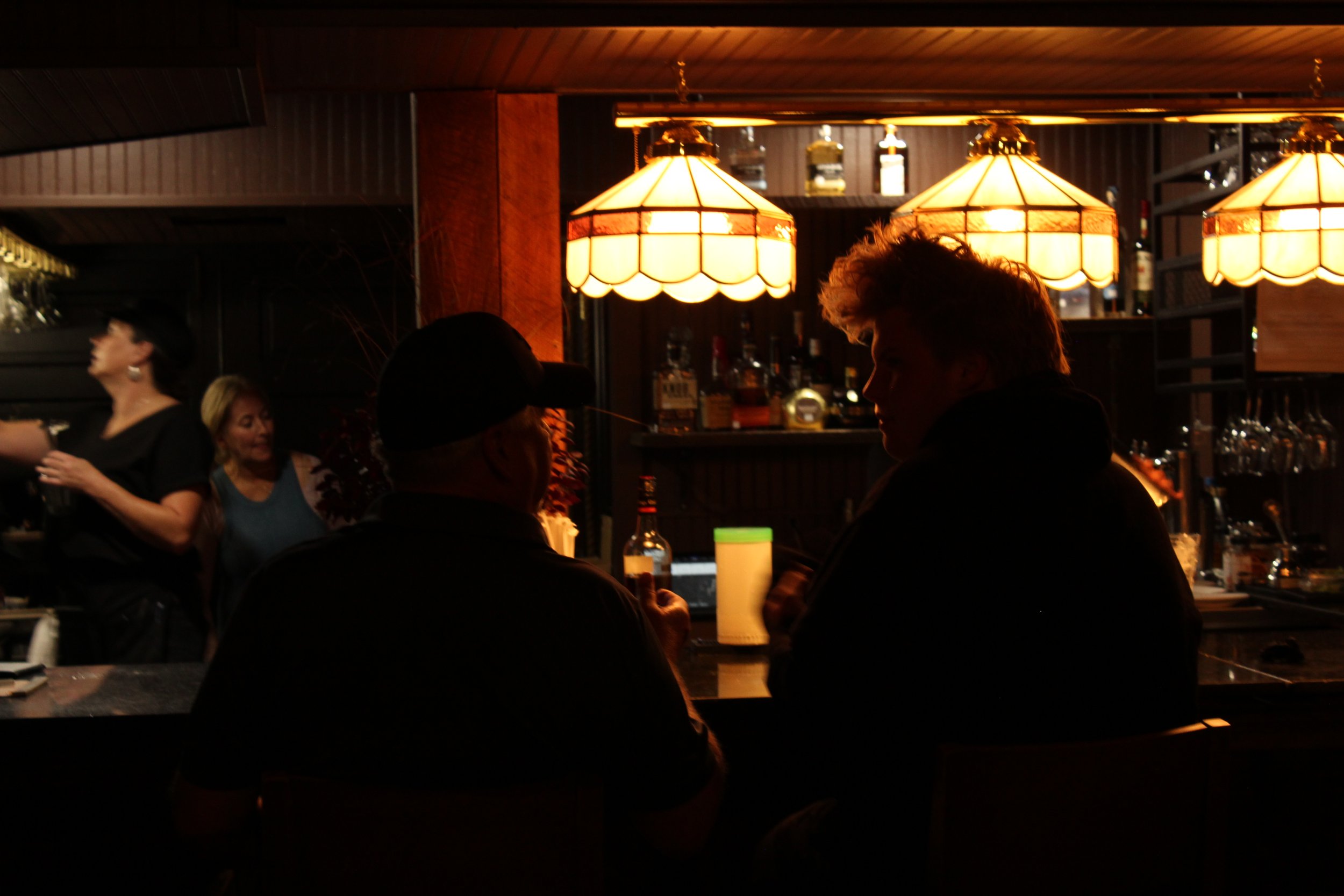People socializing inside a dimly lit bar or restaurant with warm hanging lights and shelves of bottles in the background.
