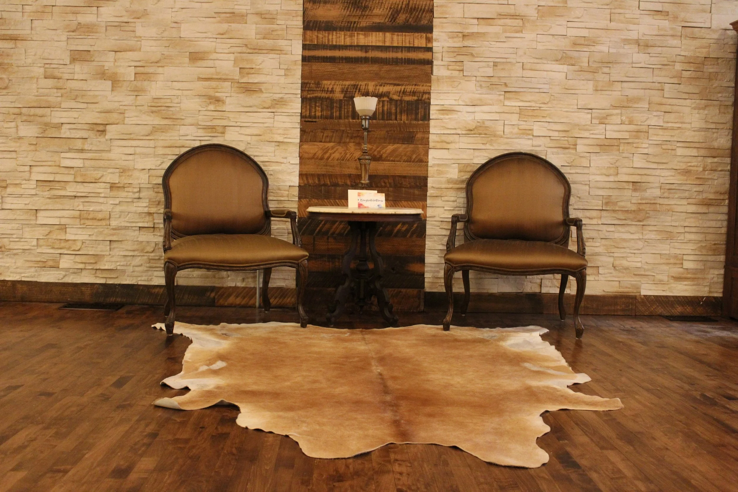 Two brown chairs with wooden frames flanking a small table with a lamp and a card that says 'Congratulations'. A cowhide rug is on the wooden floor, and the backdrop is a beige brick wall with a wooden panel in the center.