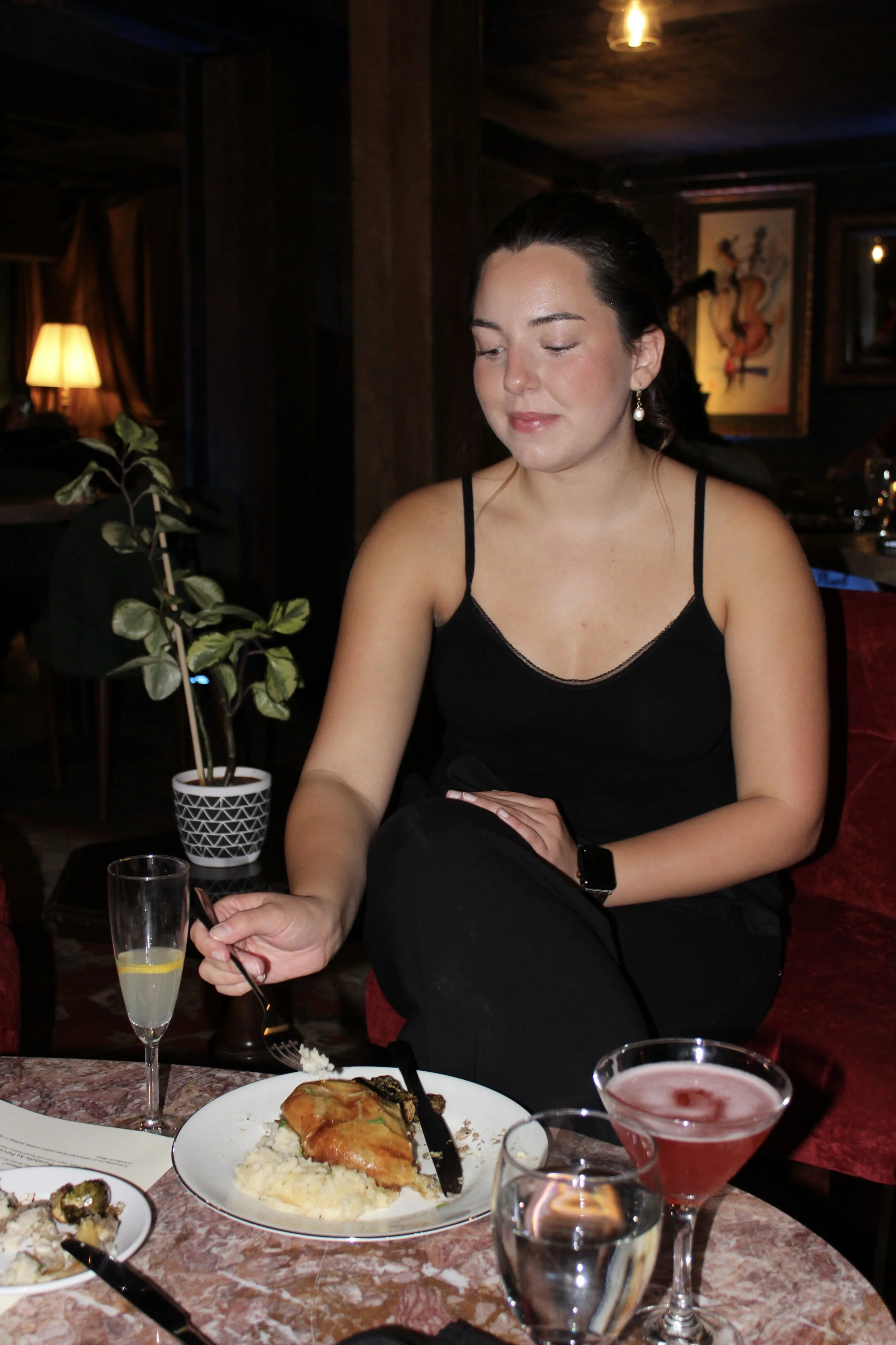 A woman with dark hair, dressed in a black top and wearing pearl earrings, is sitting at a restaurant table. She appears to be looking down and smiling slightly. The table has a plate with baked chicken, mashed potatoes, and vegetables, along with se