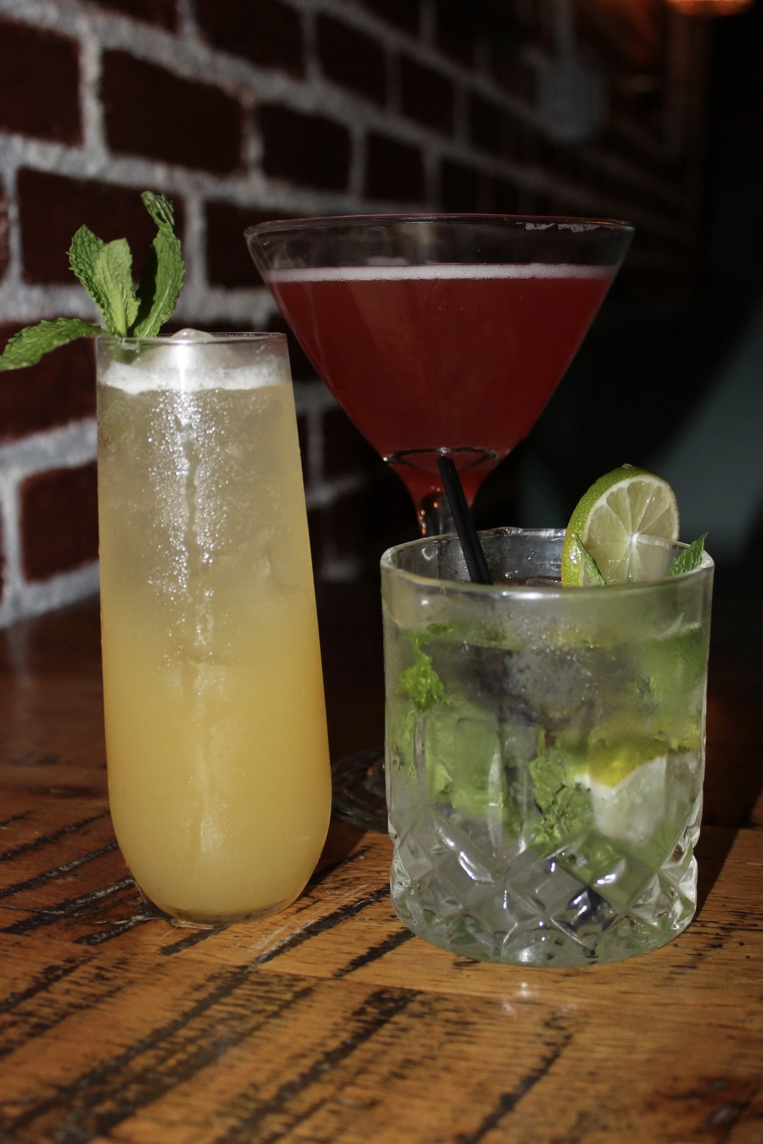 Three different cocktails placed on a wooden table with a brick wall background. The first cocktail is a tall, frothy, pale yellow drink with mint leaves as garnish. The second is a large martini glass filled with a red drink. The third is a short gl
