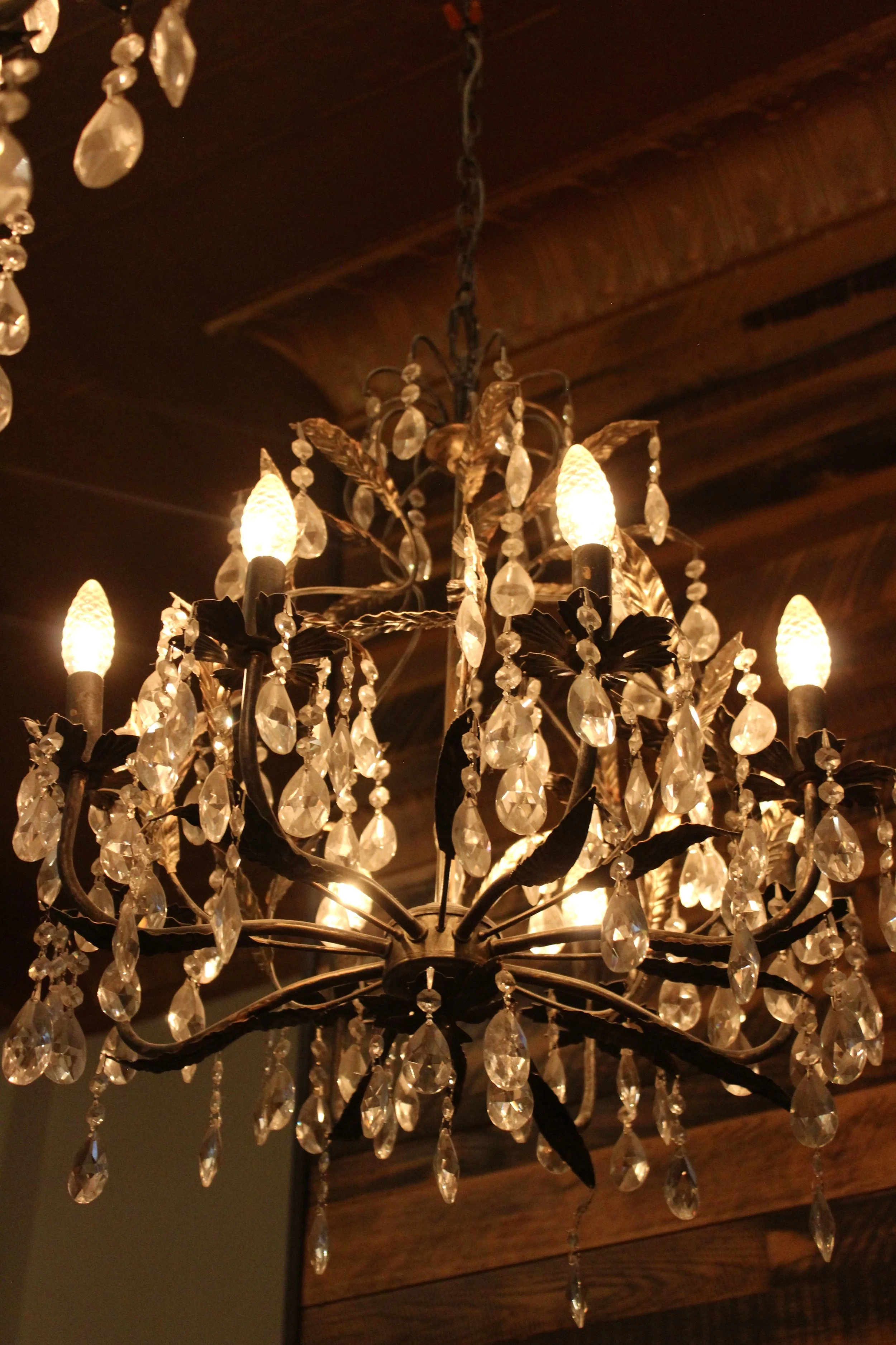 Close-up of an ornate chandelier with crystal drops hanging from dark metal arms, illuminated by candle-shaped light bulbs, against a dark wooden ceiling.