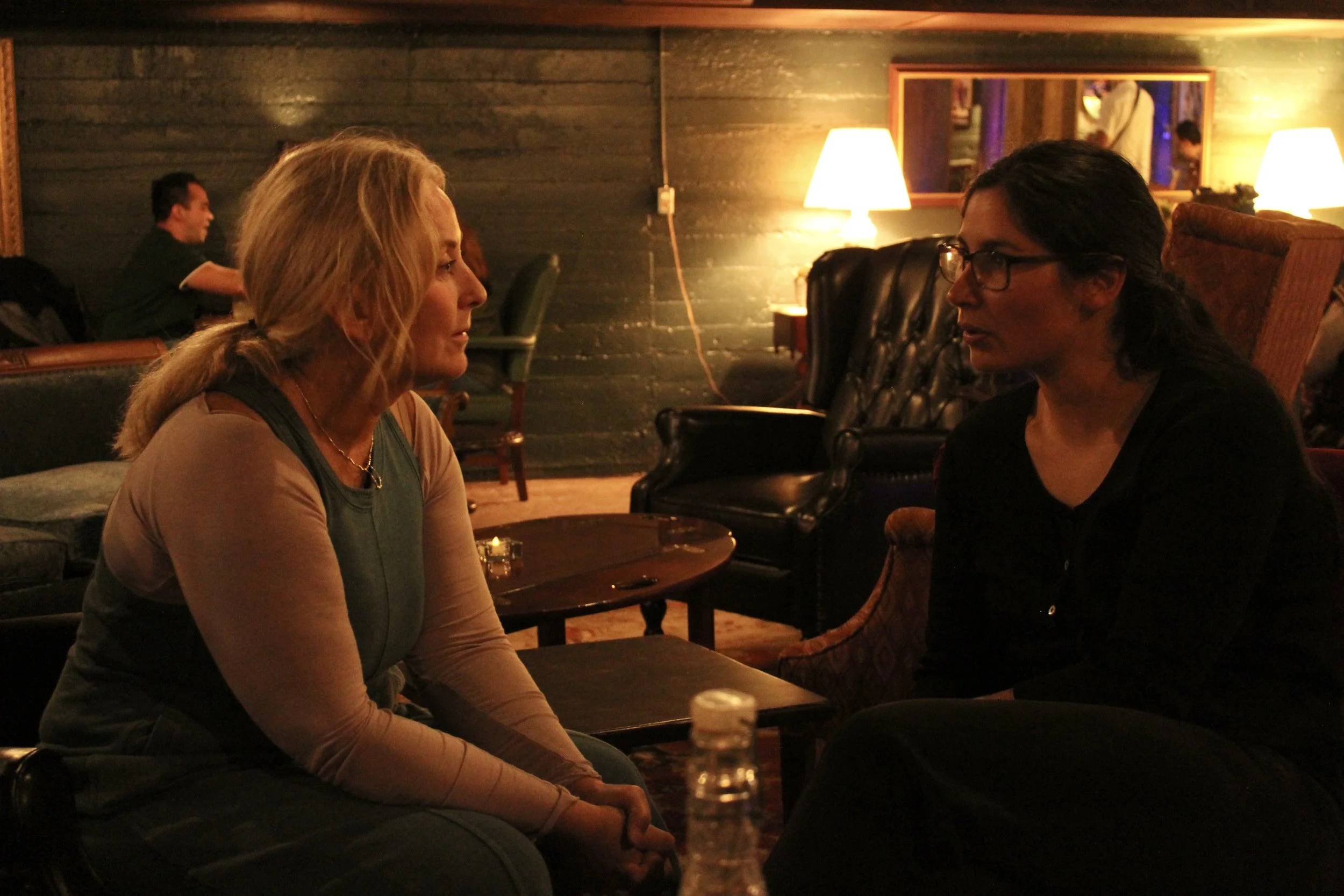 Two women are sitting face to face in a dimly lit room, engaged in conversation. There are lamps on the walls providing warm light, and additional patrons are visible in the background.