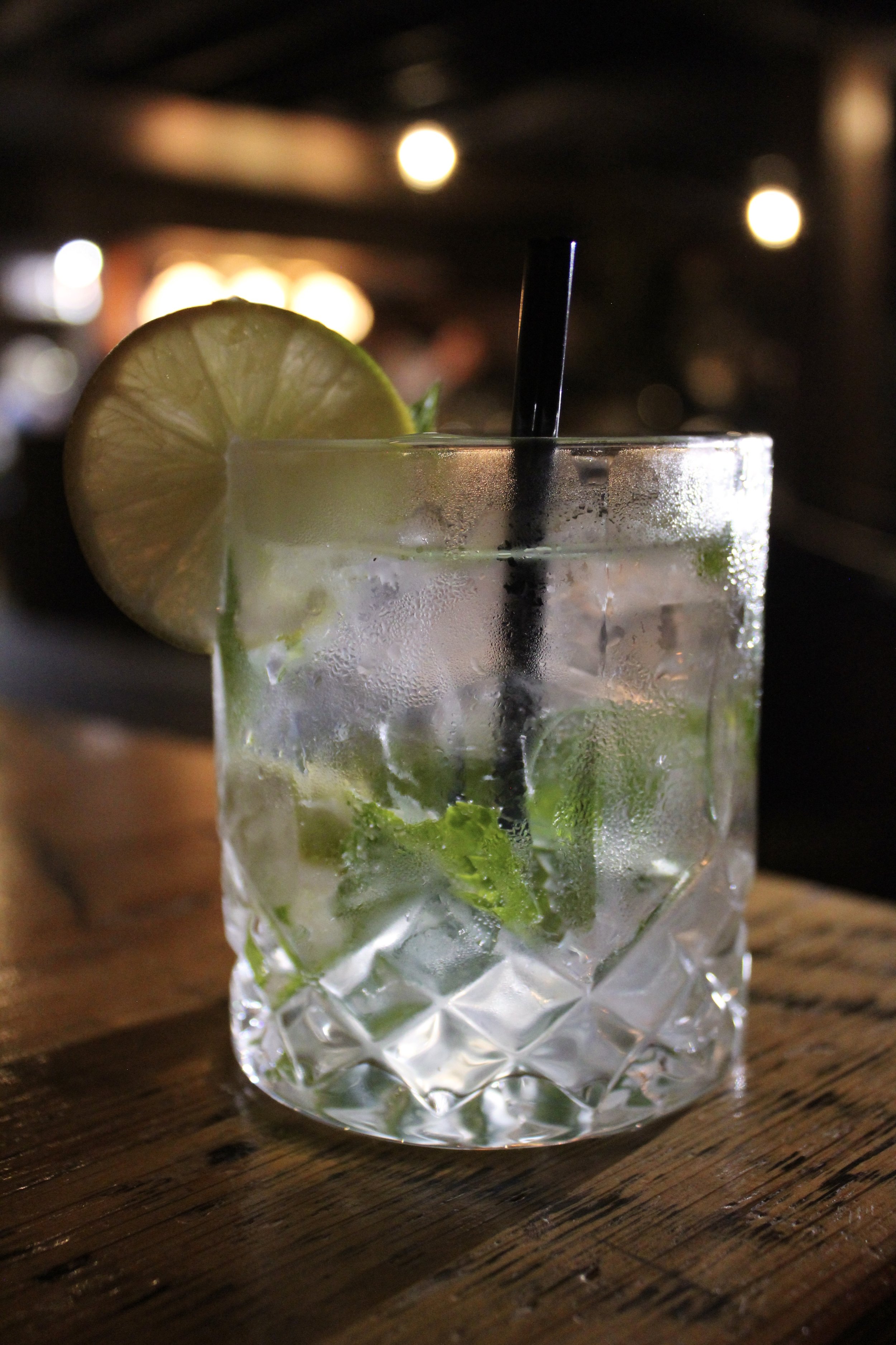 A close-up of a glass of mojito with a slice of lime on the rim, mint leaves inside, and condensation on the outside, placed on a wooden surface in a dimly lit setting.