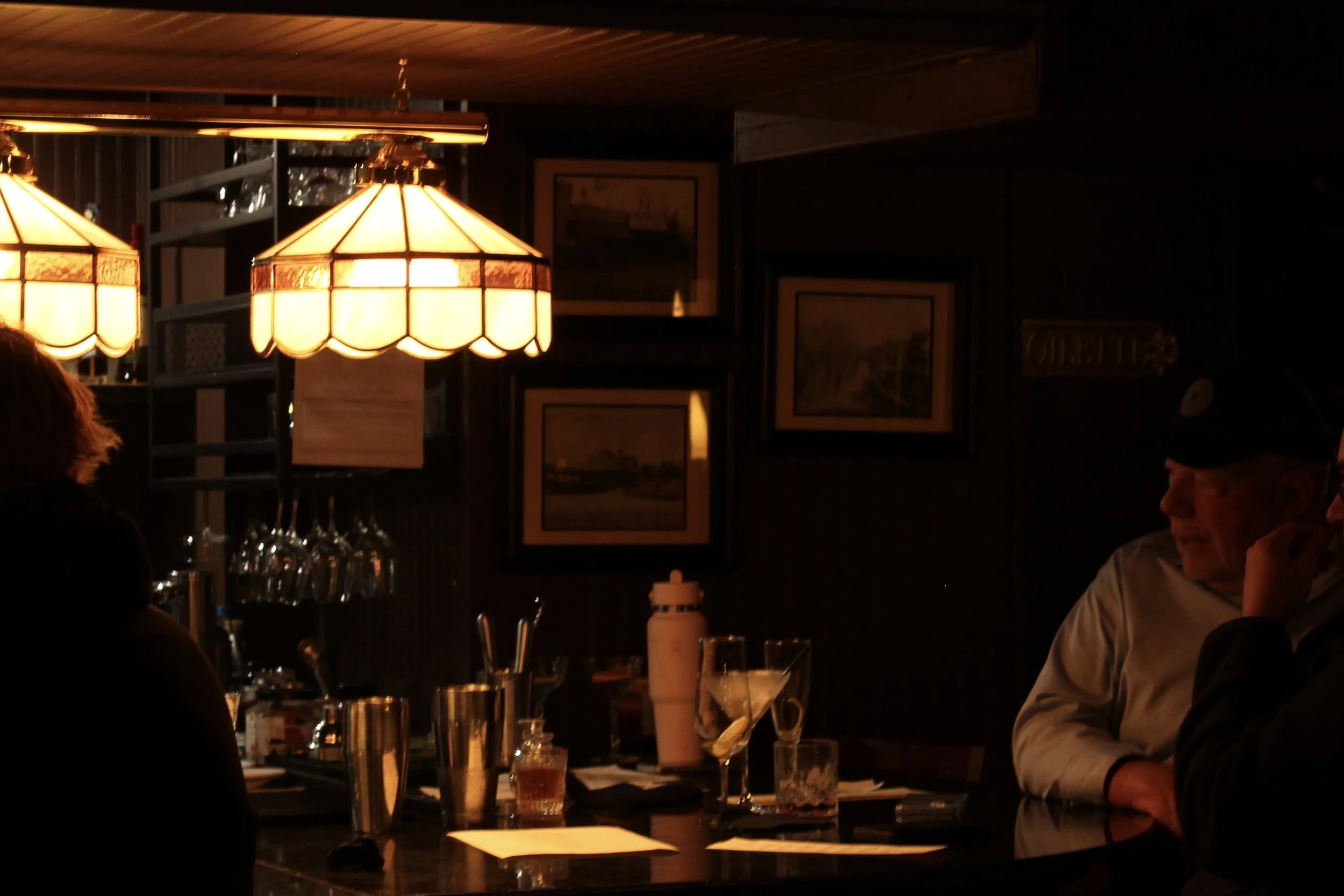 A cozy restaurant interior with dim lighting, hanging lamps, and framed pictures on the wall. People are seated at the bar, enjoying drinks and conversation.