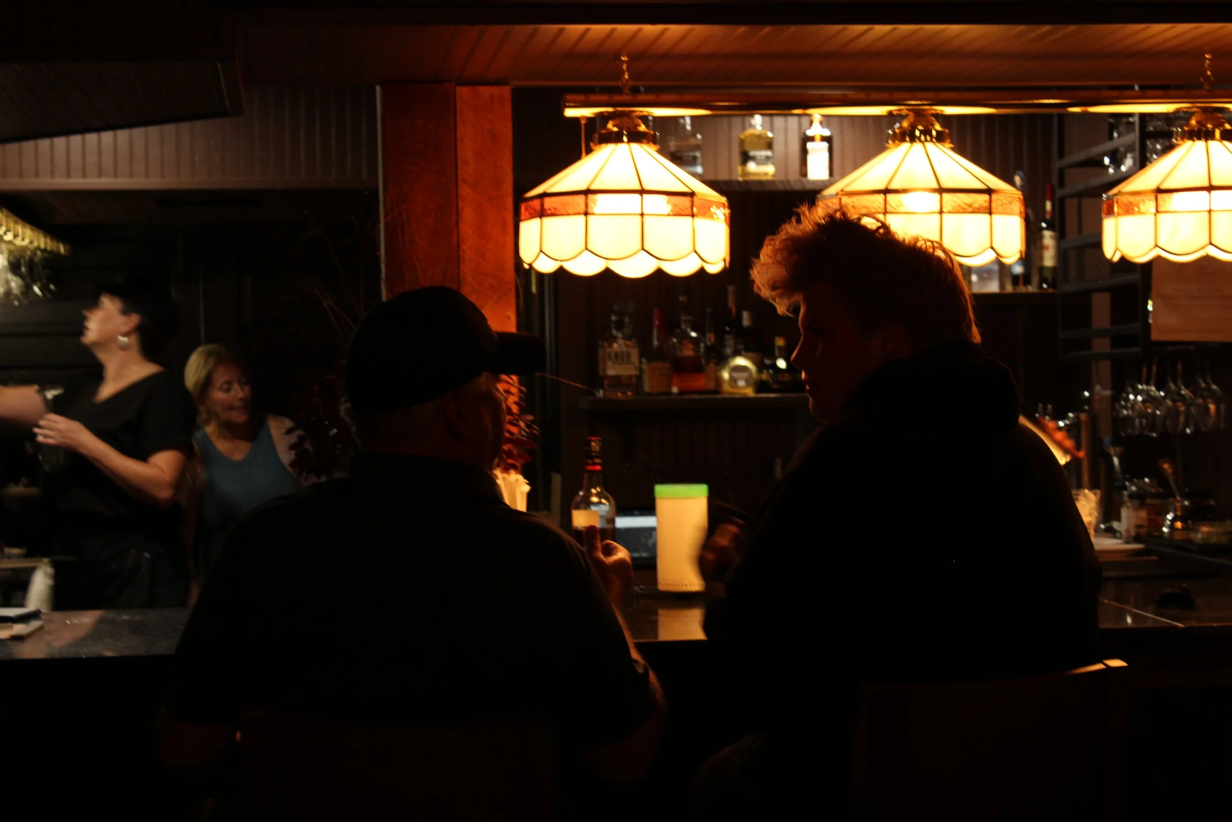 Dimly lit bar scene with three large hanging lamps, several patrons engaging in conversations, and shelves stocked with bottles in the background.