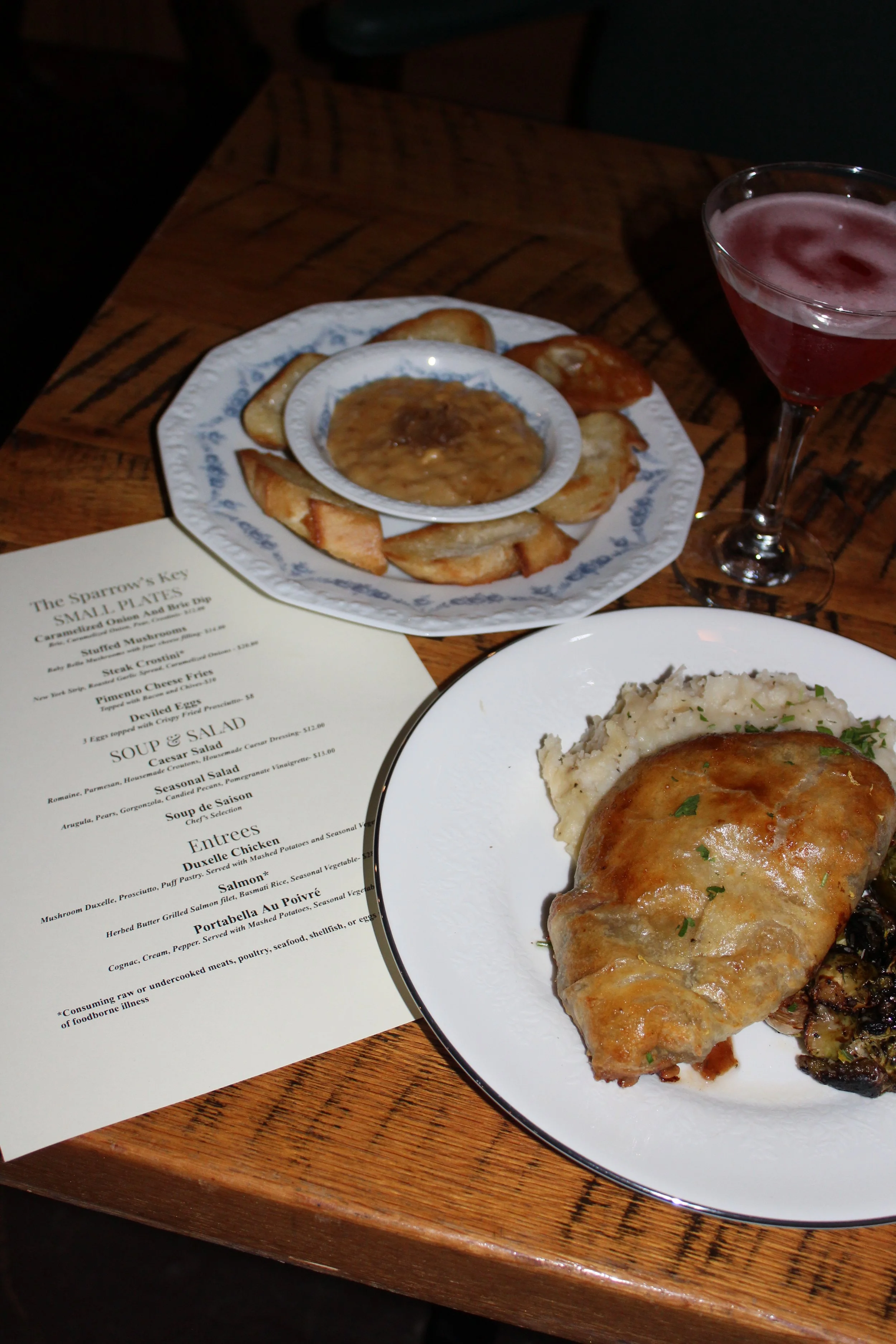A table with a plate of baked chicken breast topped with sauce, mashed potatoes with gravy, and a small bowl of gravy or sauce, along with a drink in a martini glass, a menu, and a second plate of food in the foreground.