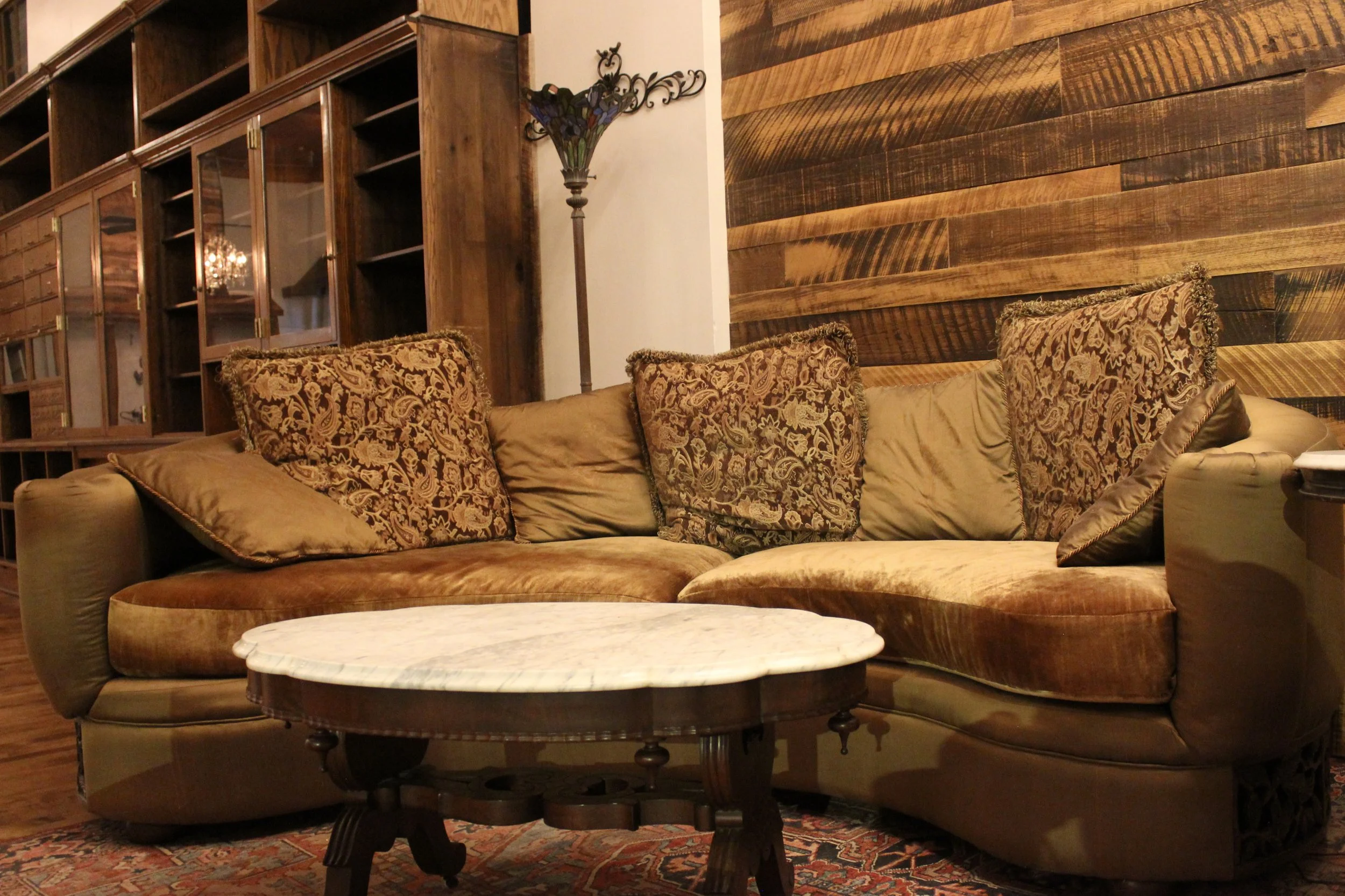 A cozy living room with a large, curved beige sofa decorated with patterned cushions. There's a round marble-topped coffee table in front of the sofa. The background features a wooden wall panel and a tall lamp with a floral-shaped stained glass lamp