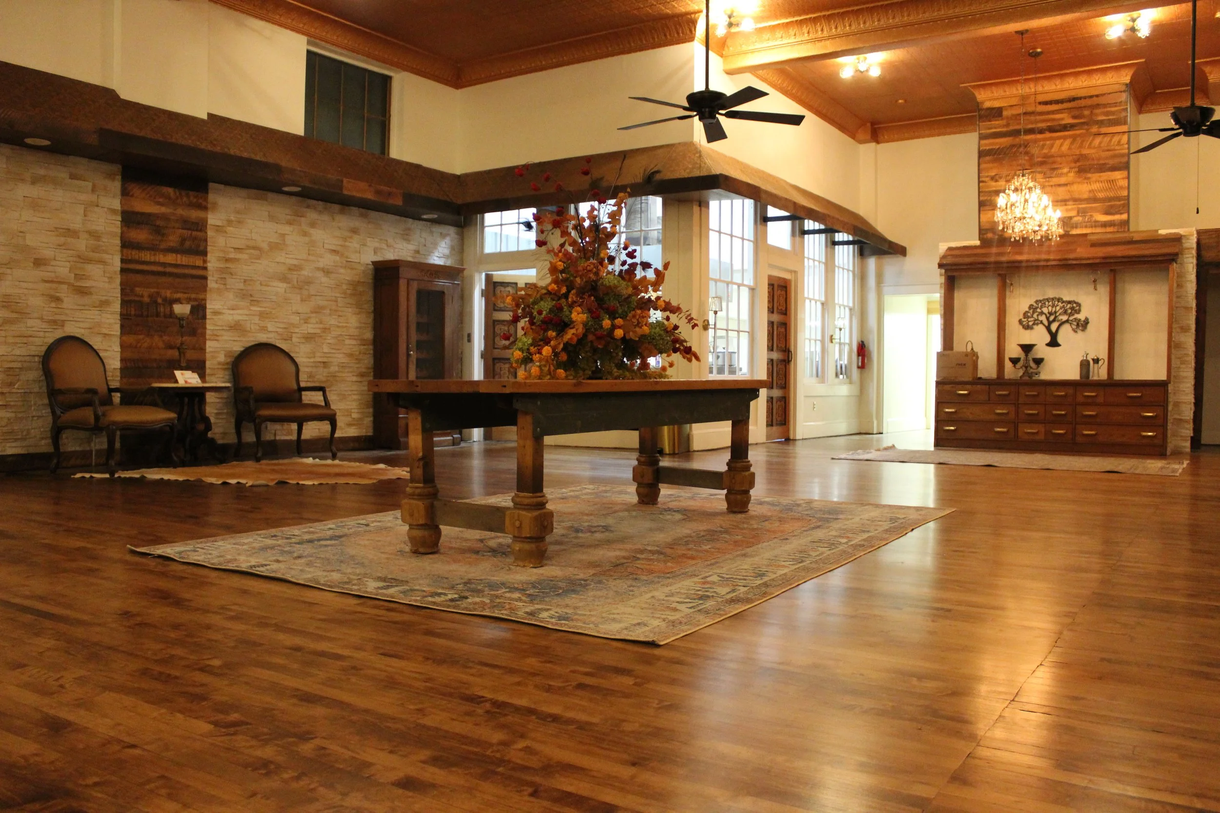 Interior of a spacious room with a high wooden ceiling, a chandelier, and a large floral arrangement on a rustic wooden table. There are two ceiling fans, a stone and wood accent wall, and a pair of chairs against the wall.