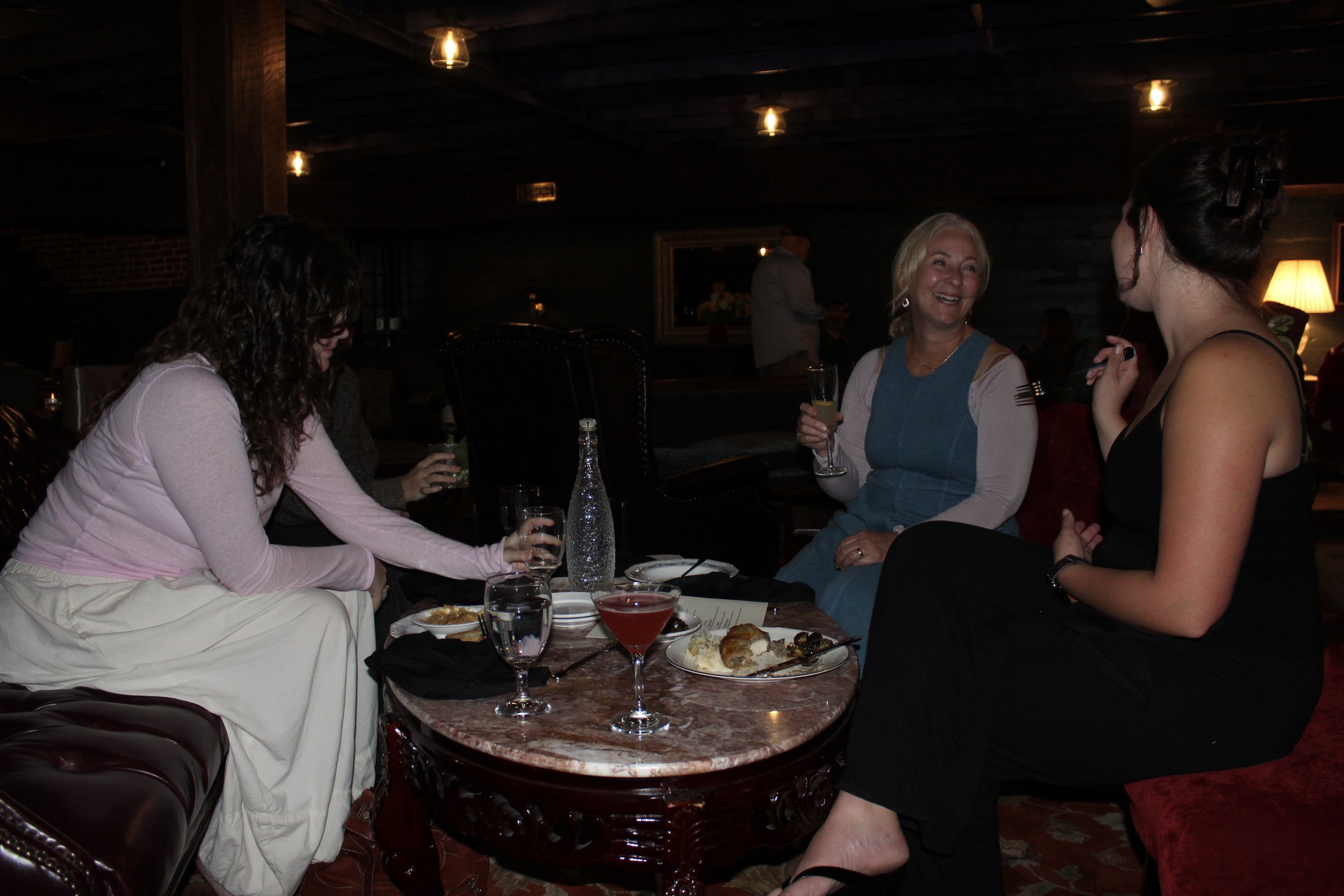 Four women sitting around a table, engaged in conversation at a gathering or party, with food and drinks on the table in a dimly lit room.