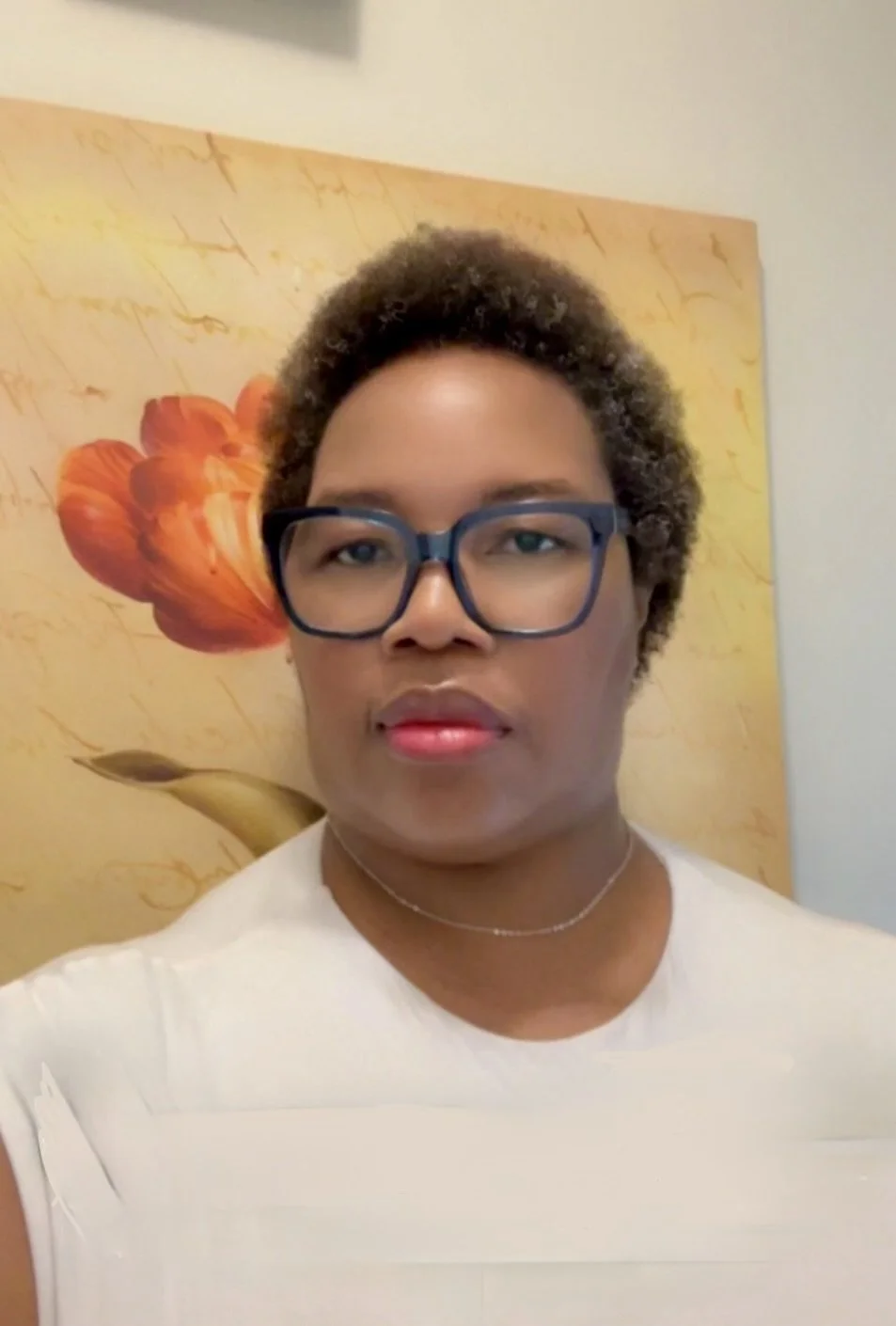 A woman with short, curly hair and glasses, wearing a light-colored top and a necklace, standing in front of a colorful flower-themed background.