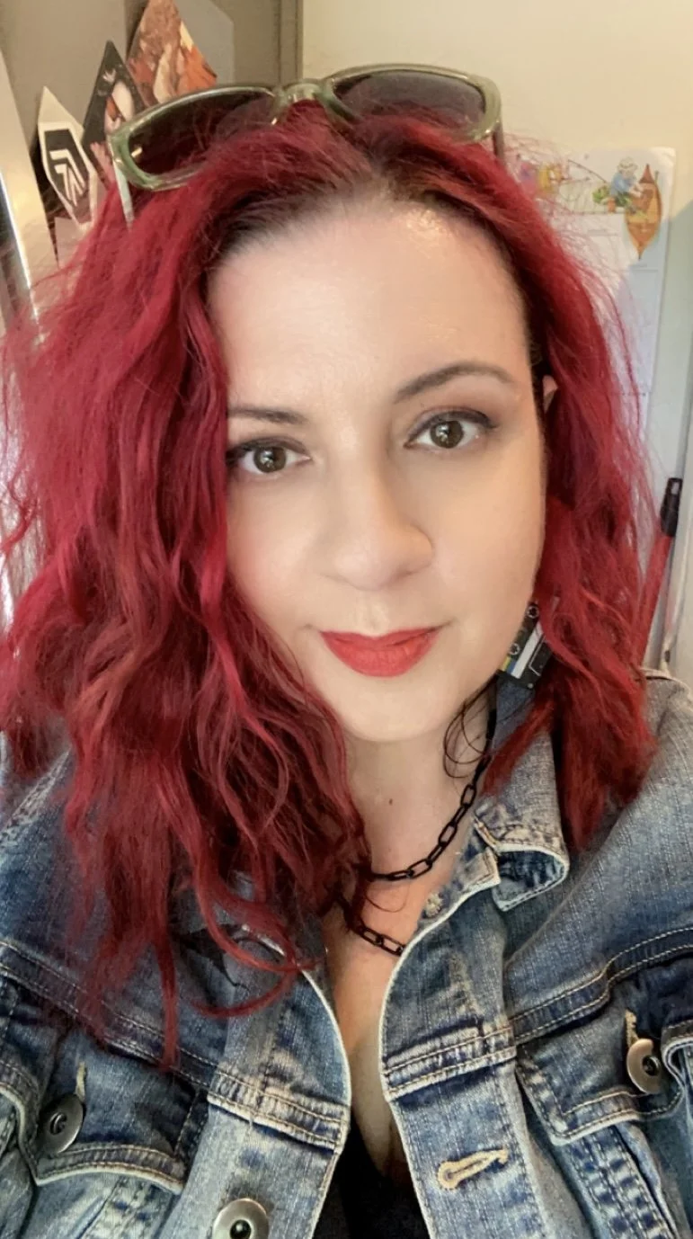 A woman with vibrant red wavy hair, wearing sunglasses on her head, a denim jacket, and a black choker necklace.