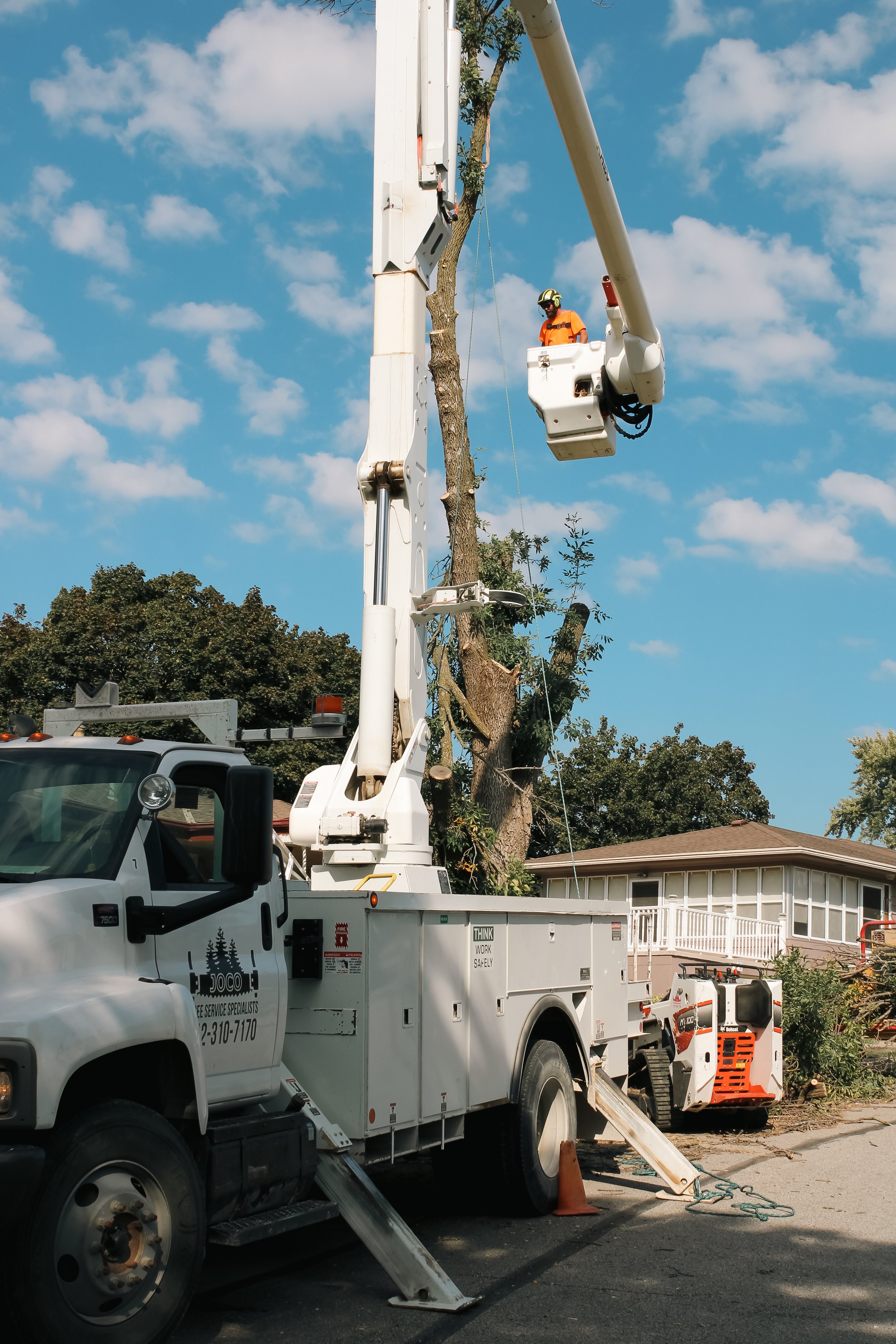 A worker in an orange uniform and helmet in a bucket lift trimming a tall tree with fallen branches and debris nearby on a residential street.