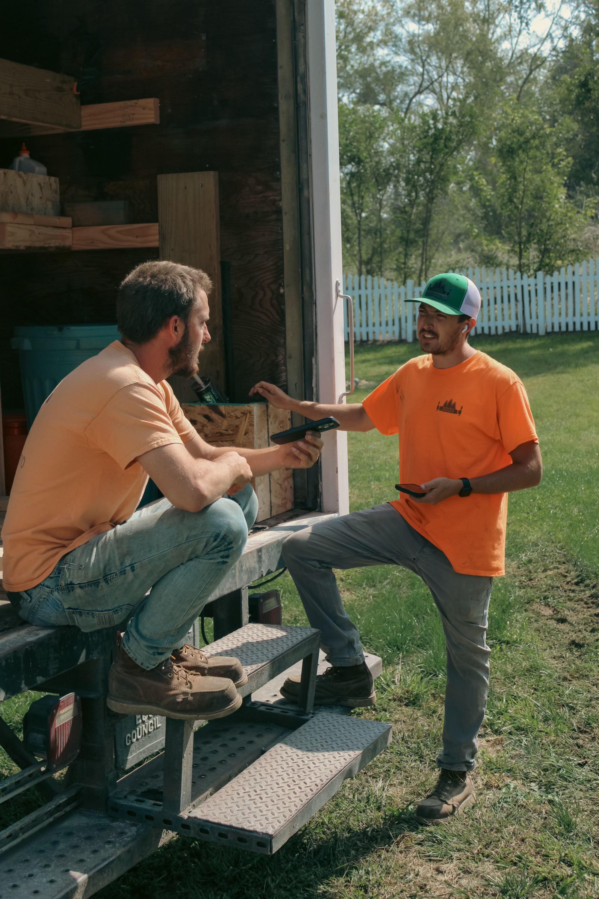 Two men having a conversation outside near a wooden shed, one sitting on the steps and the other standing, holding a mobile phone, with green grass and a white picket fence in the background.