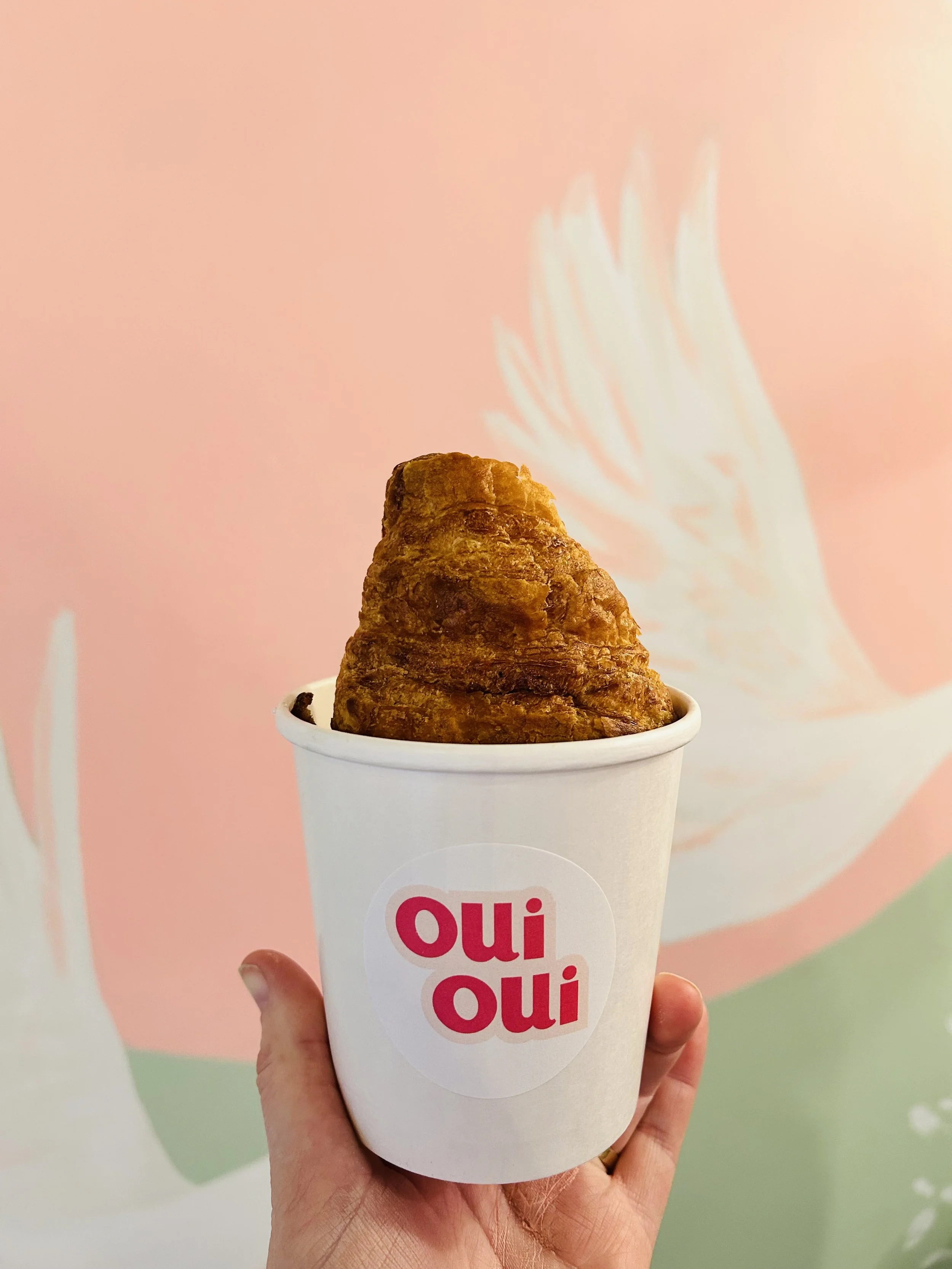 A hand holding a white cup with a croissant inside, against a pink background with a white and pastel-colored silhouette of a bird.