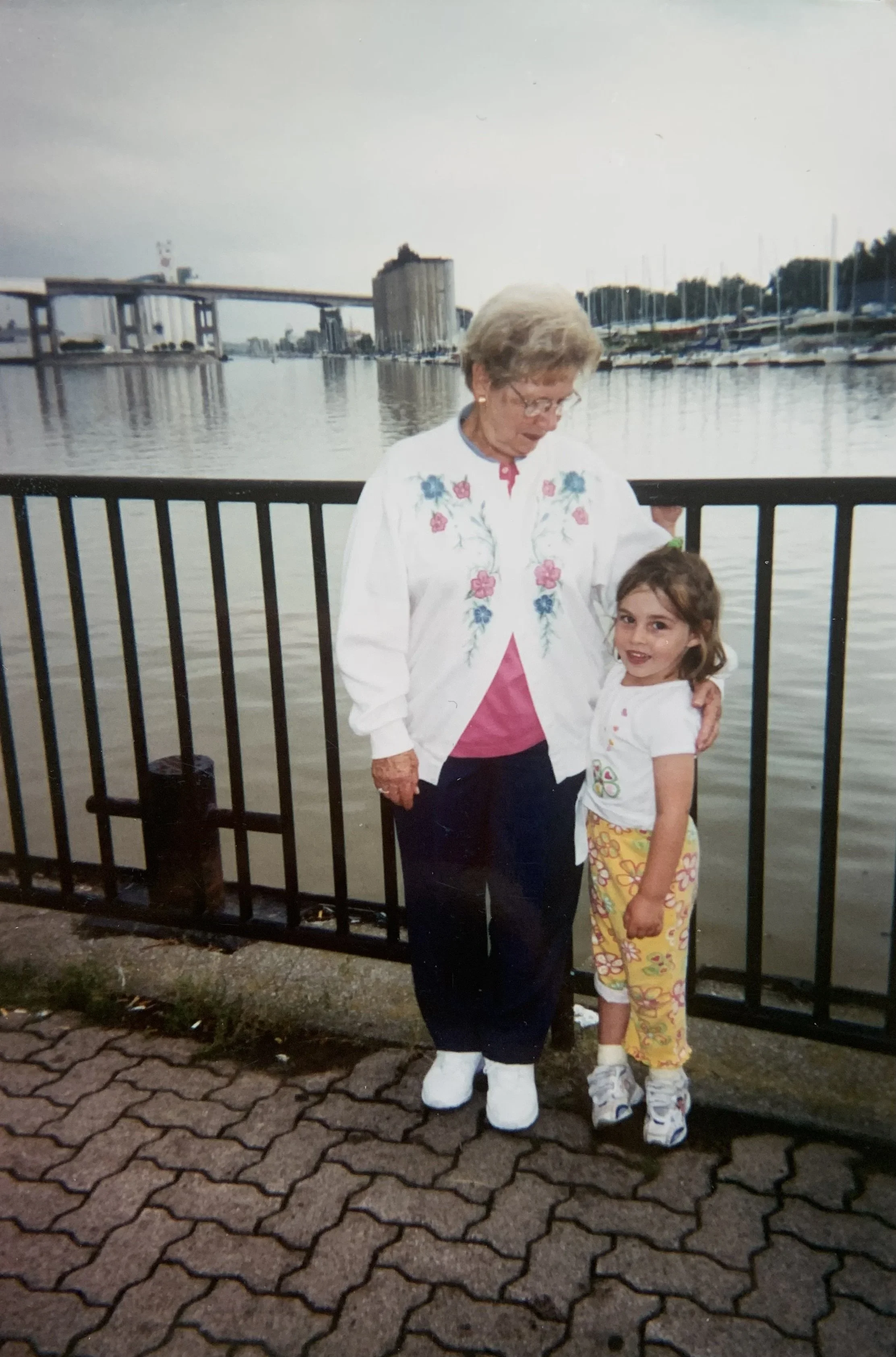 An elderly woman and young girl stand together by a waterfront, with boats and buildings in the background, holding each other and smiling.