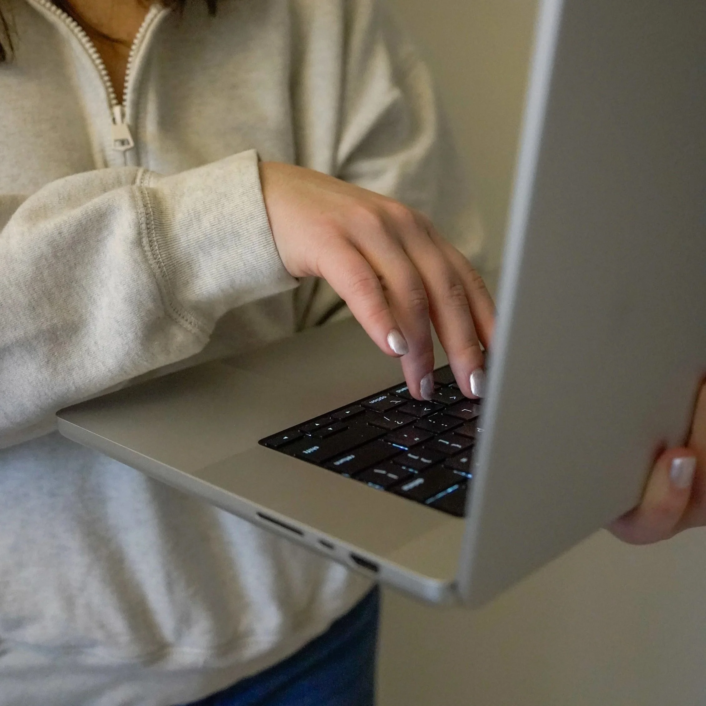 A person in a gray hoodie typing on a silver laptop with black keys.