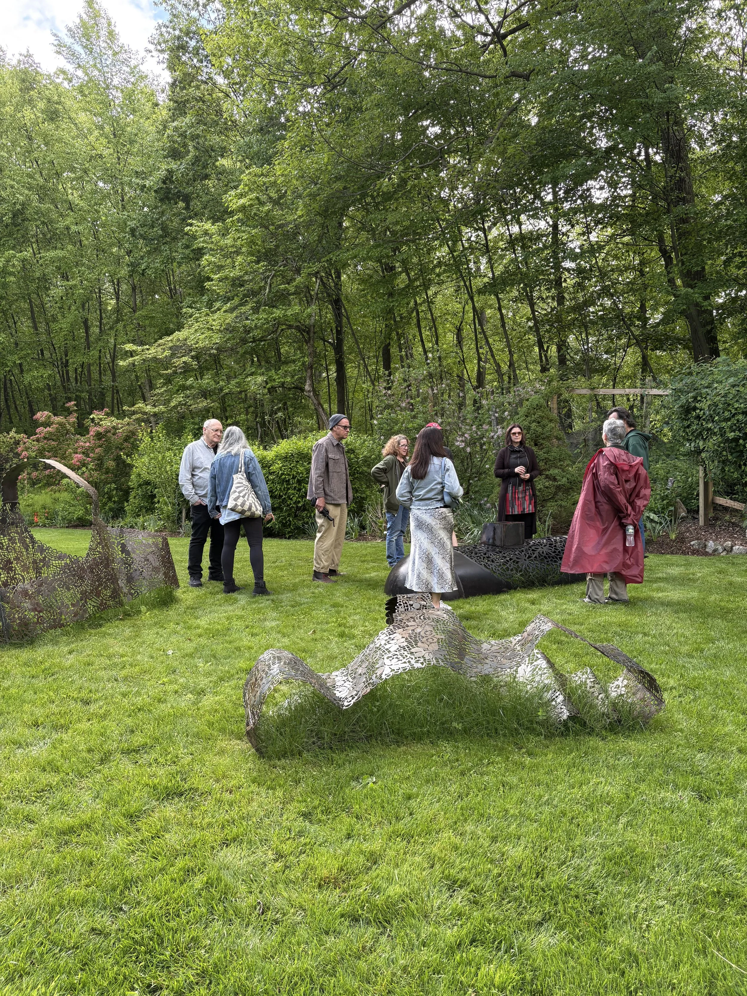 Group of people standing and talking outdoors in a lush, green garden with trees and metal sculptures in the grass.