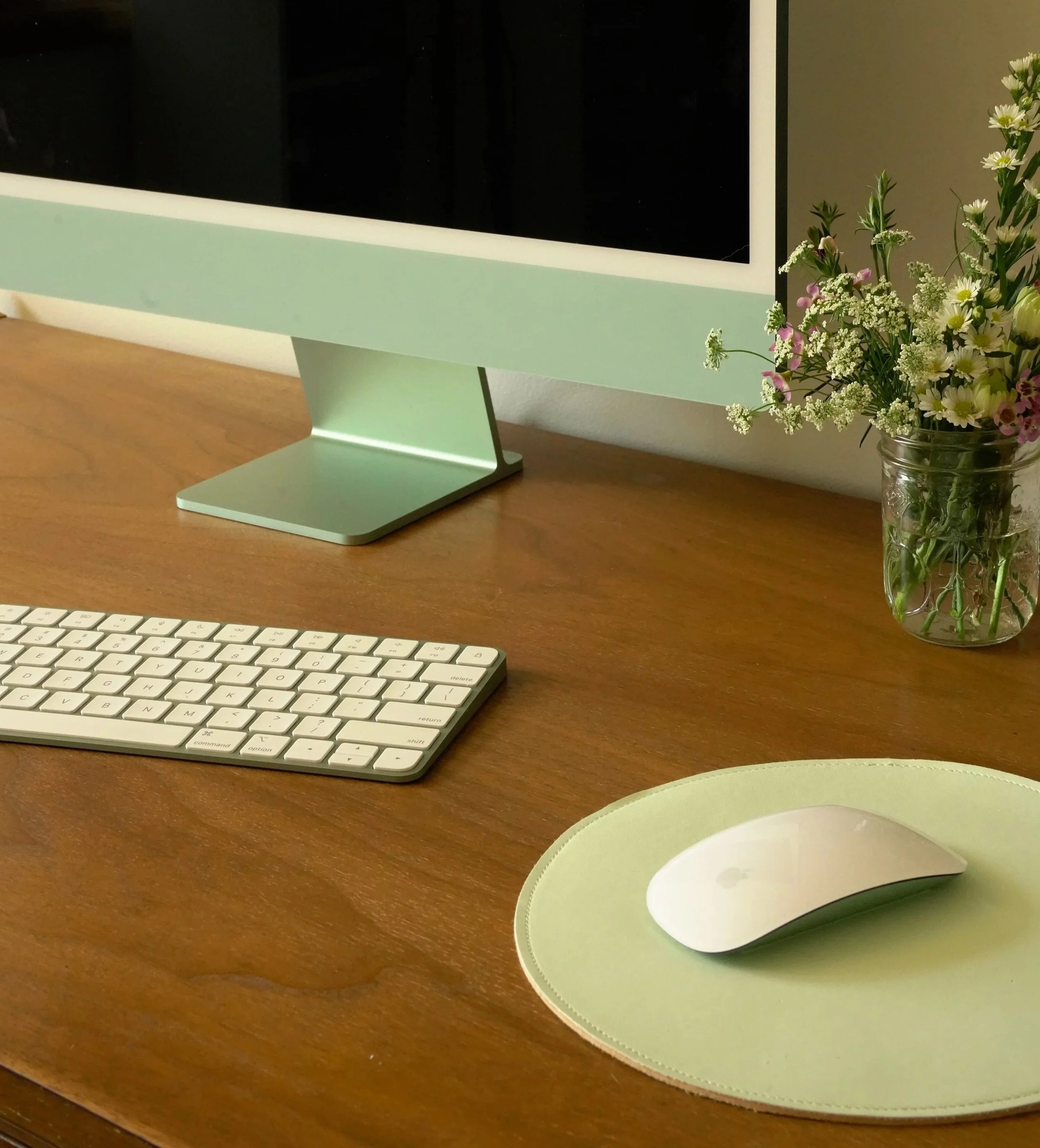 A computer monitor on a wooden desk, accompanied by a wireless keyboard, a wireless mouse on a green mouse pad, and a small glass vase with a bouquet of white and pink flowers.