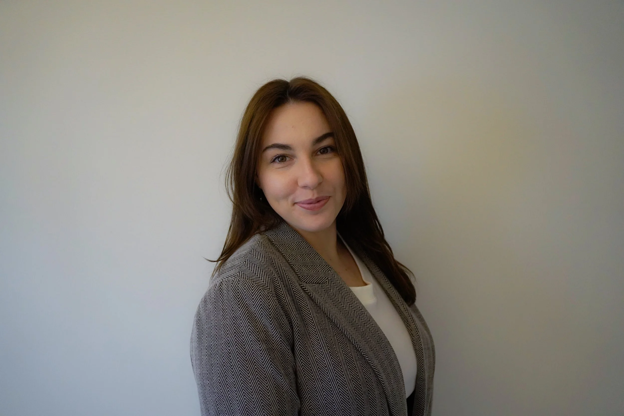 A young woman with brown hair and light skin, wearing a gray blazer over a white top, standing against a plain light-colored wall