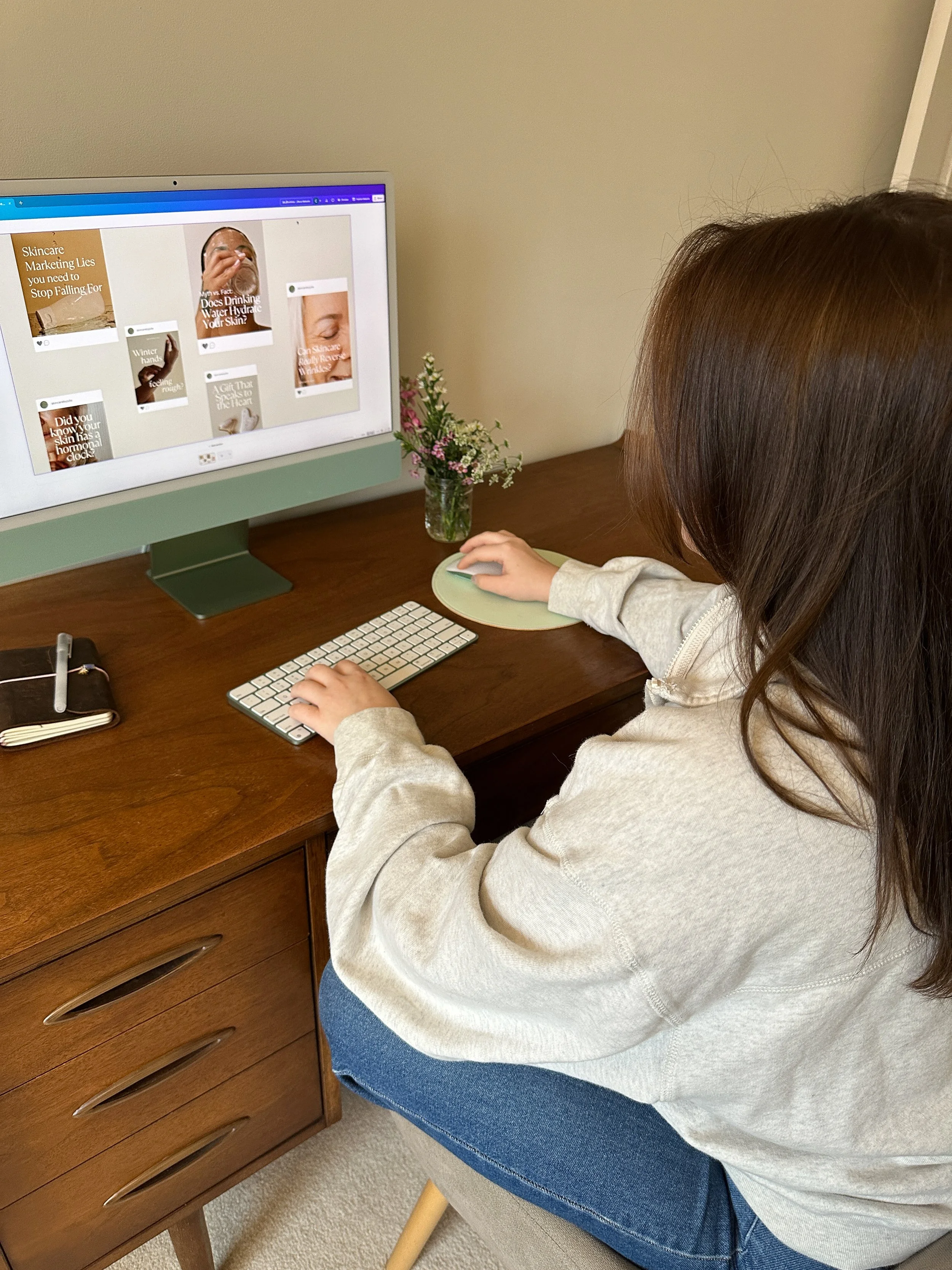 A person with brown hair using a desktop computer on a wooden desk. The monitor displays social media posts or articles about skincare and hydration. There is a small vase with flowers, a notebook, and a pen on the desk.